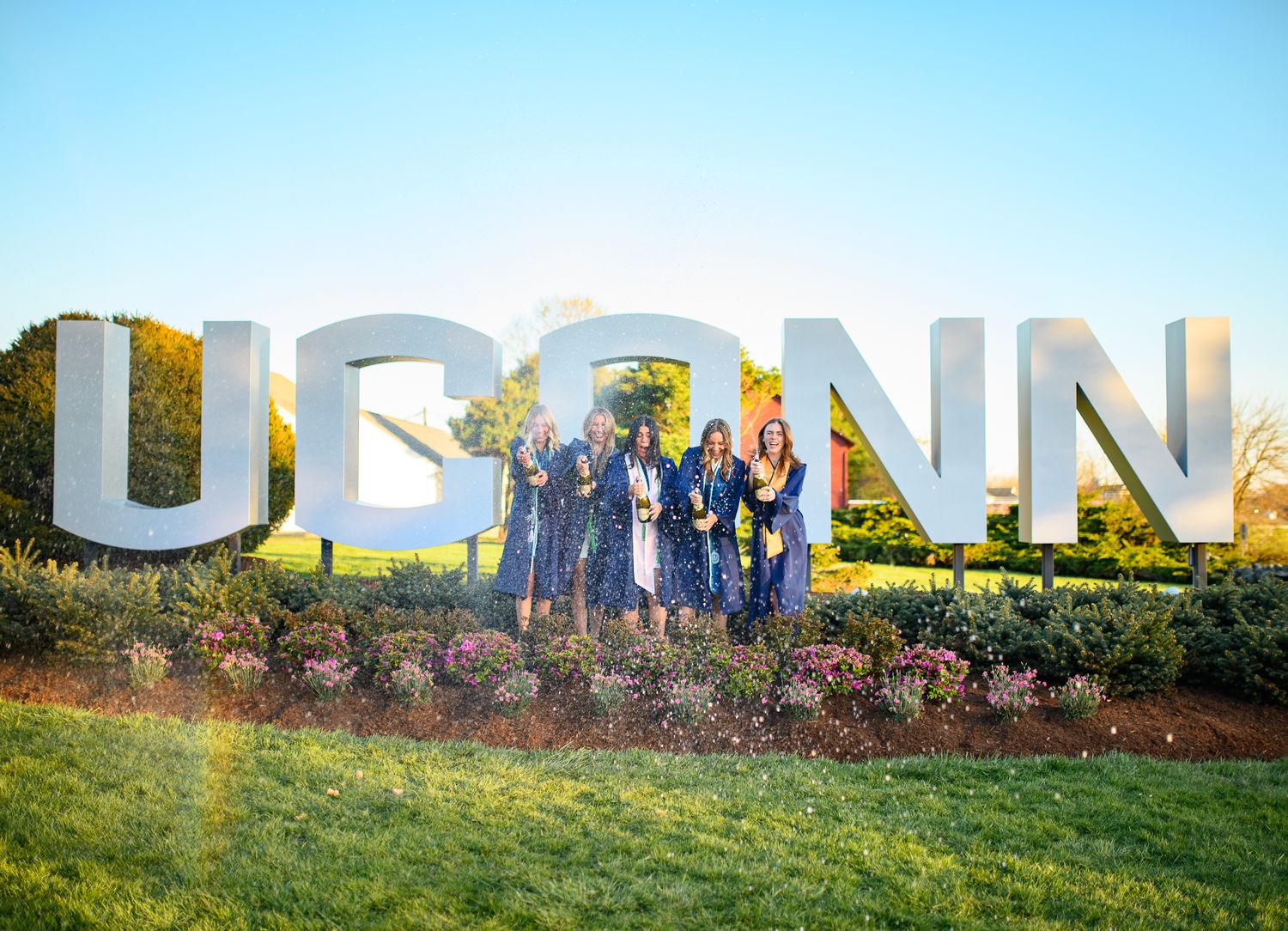 Graduates in blue caps and gowns celebrate in front of large UCONN letters on a sunny day.