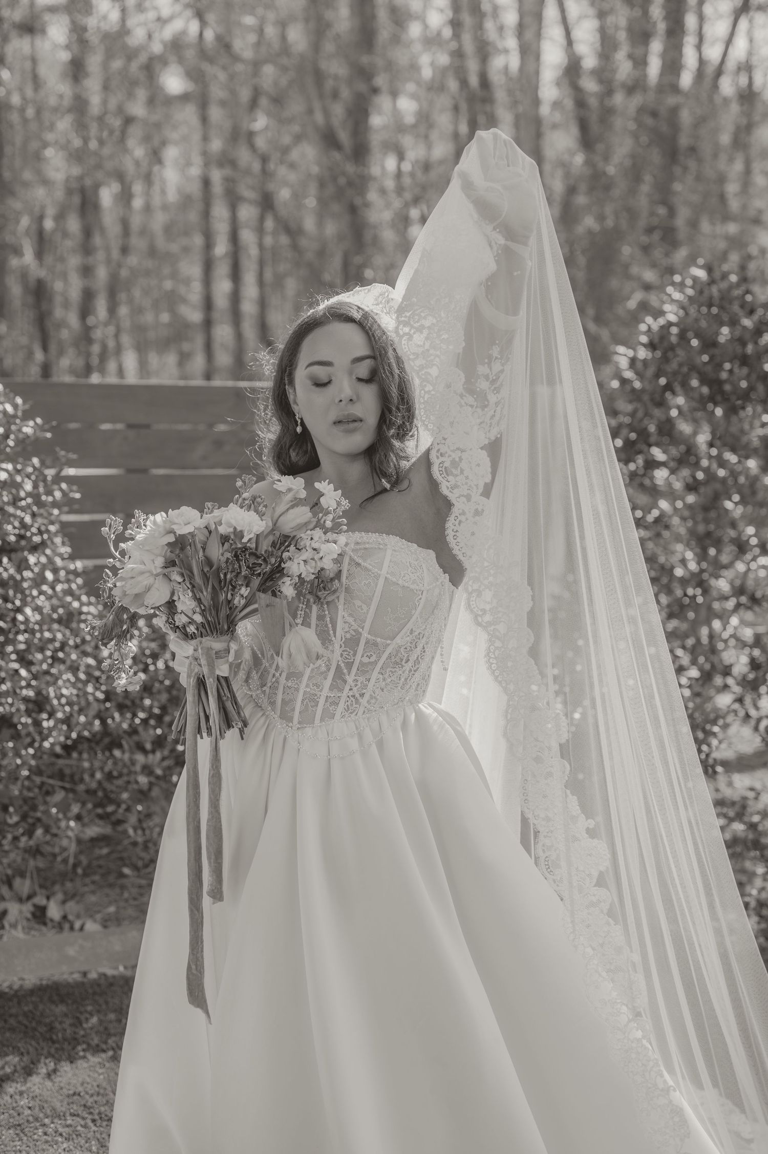 Black and white wedding photo showing a bride in a corseted ballgown with long lace veil holding a bouquet outdoors.