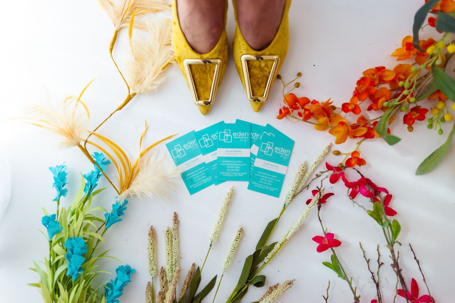 Elegant gold shoes paired with teal invitation cards and colorful flowers on a white background, styled for an event.