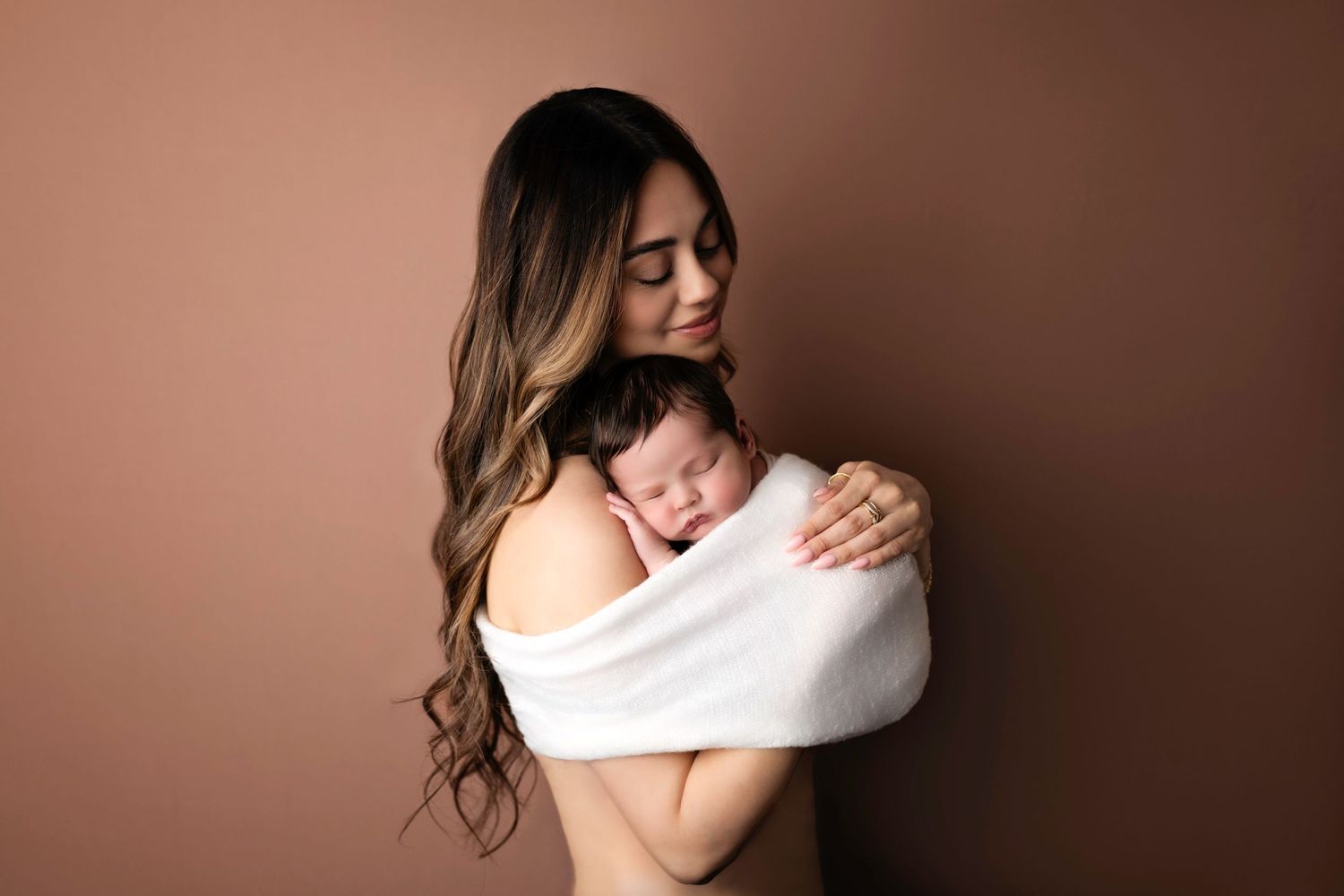 A tender moment captured as a mother in white holds her sleeping newborn against a mocha-colored backdrop.