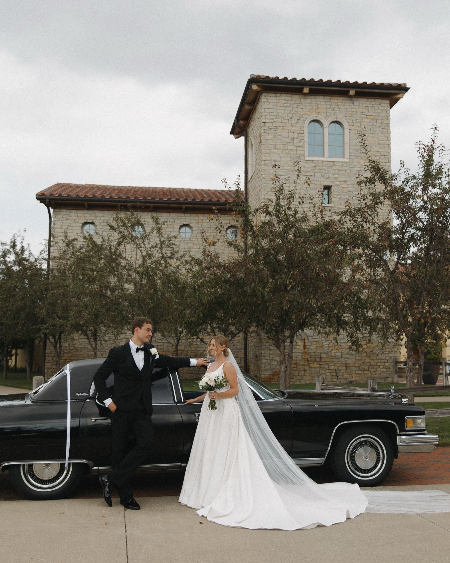 A vintage black Cadillac parked in front of a stone building with wedding couple beside it.