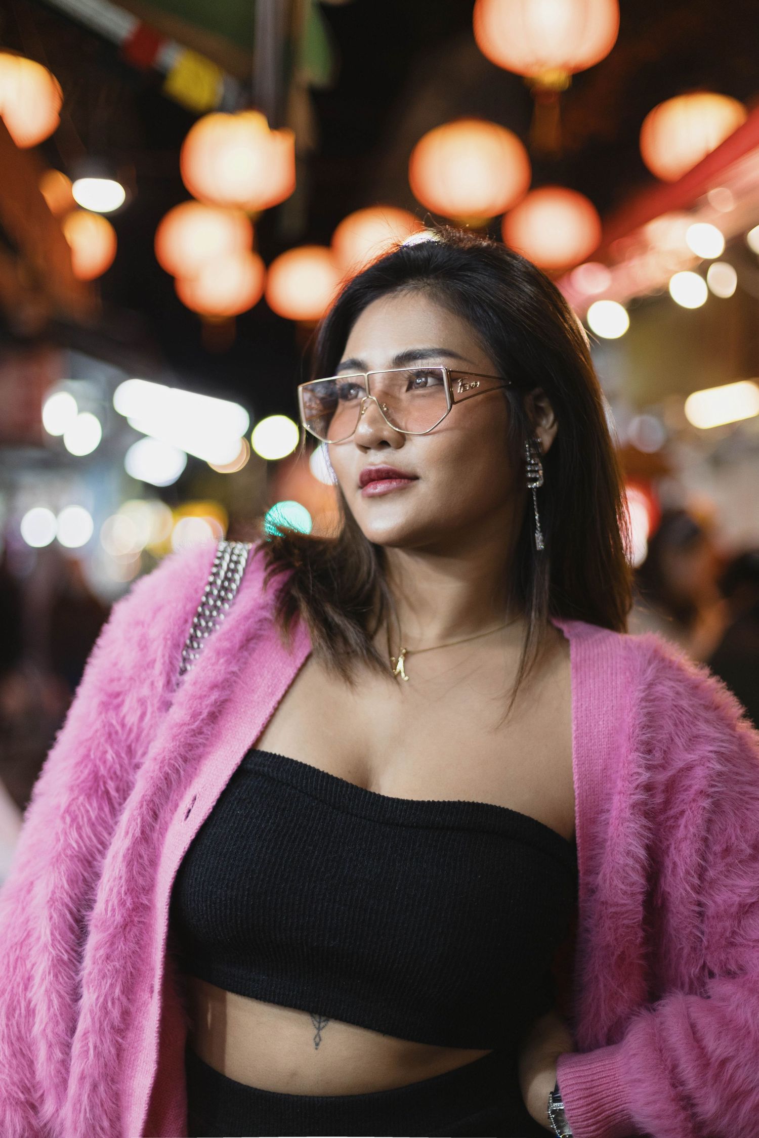Trendy woman in pink fuzzy cardigan and black crop top poses at vibrant night market with lanterns.