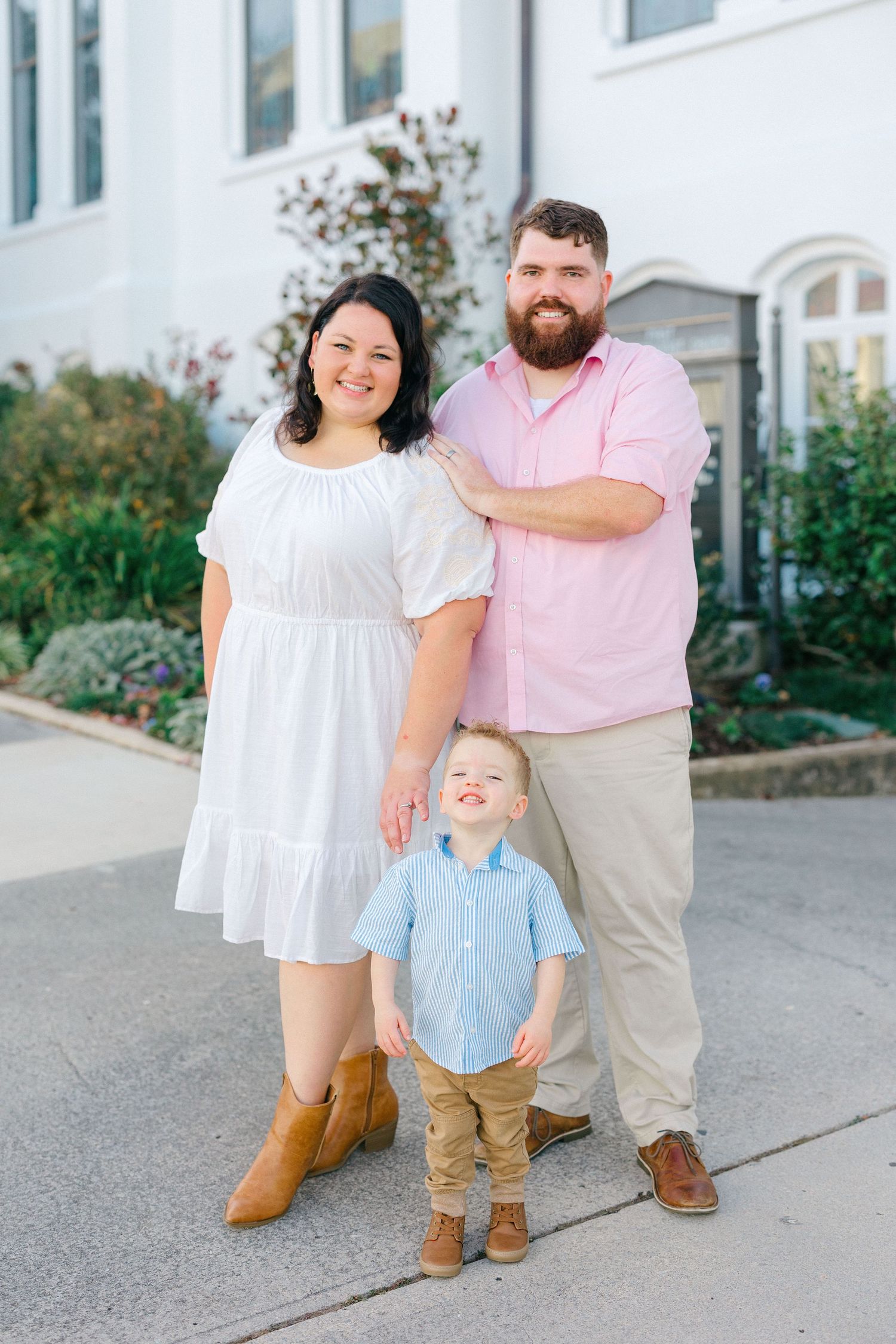 Family Photo: Mom and Dad standing with son in front of white building smiling at camera.