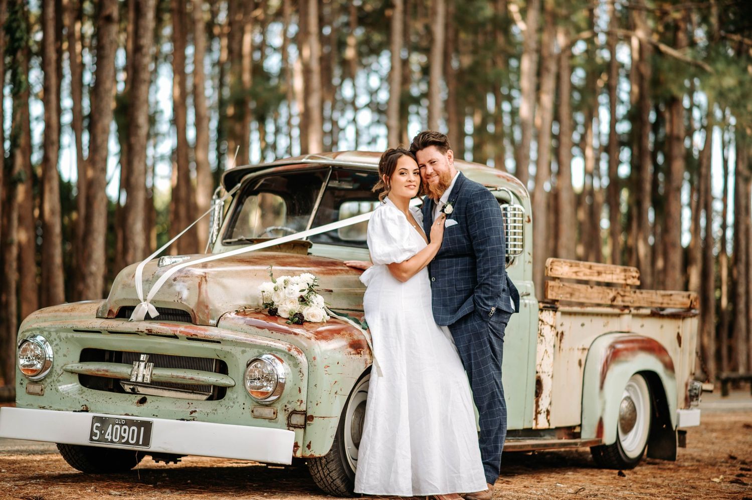 Newlyweds pose together against a rustic pickup truck surrounded by tall pine trees.