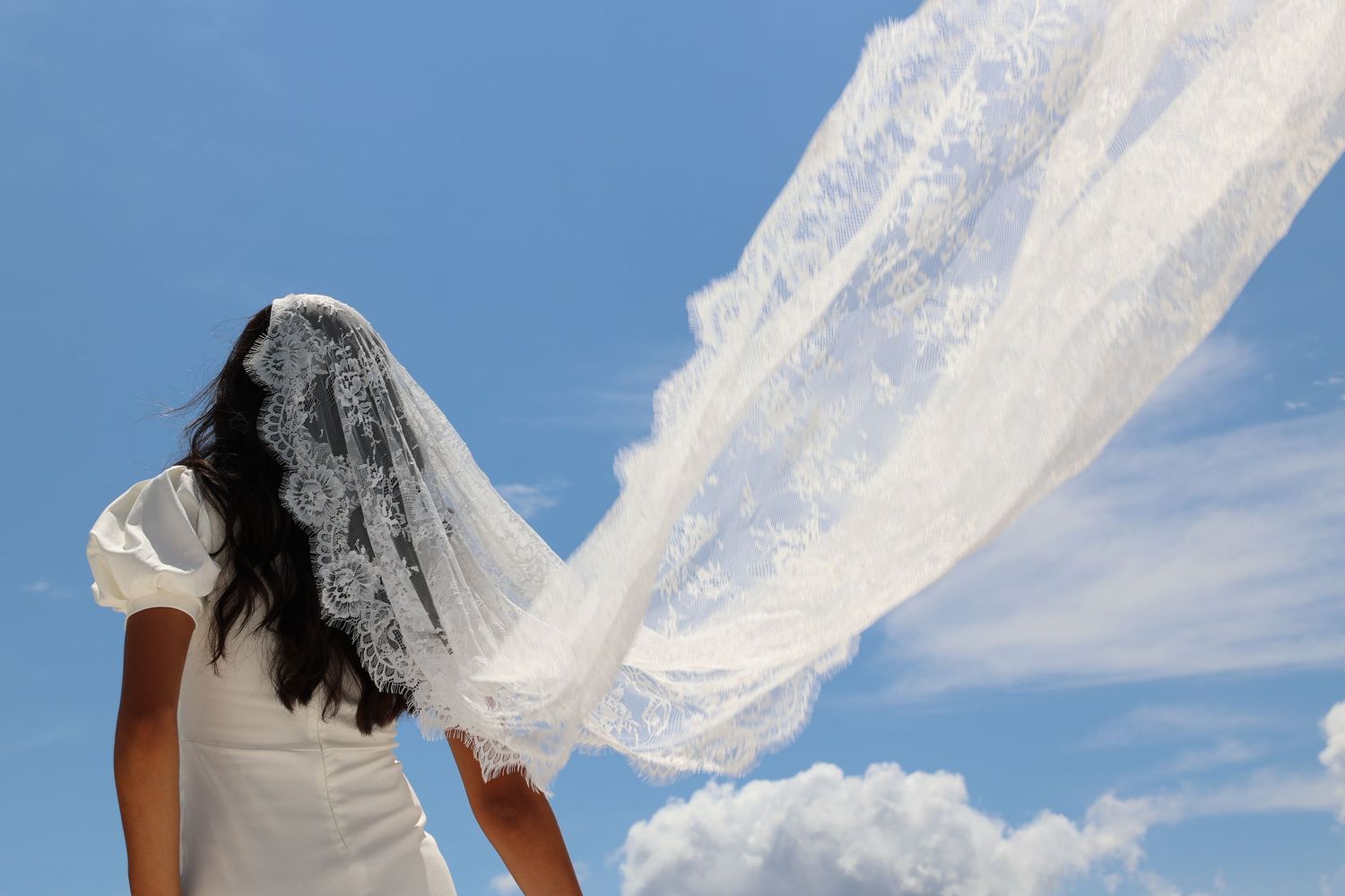 Bride's white veil flowing dramatically against blue sky.