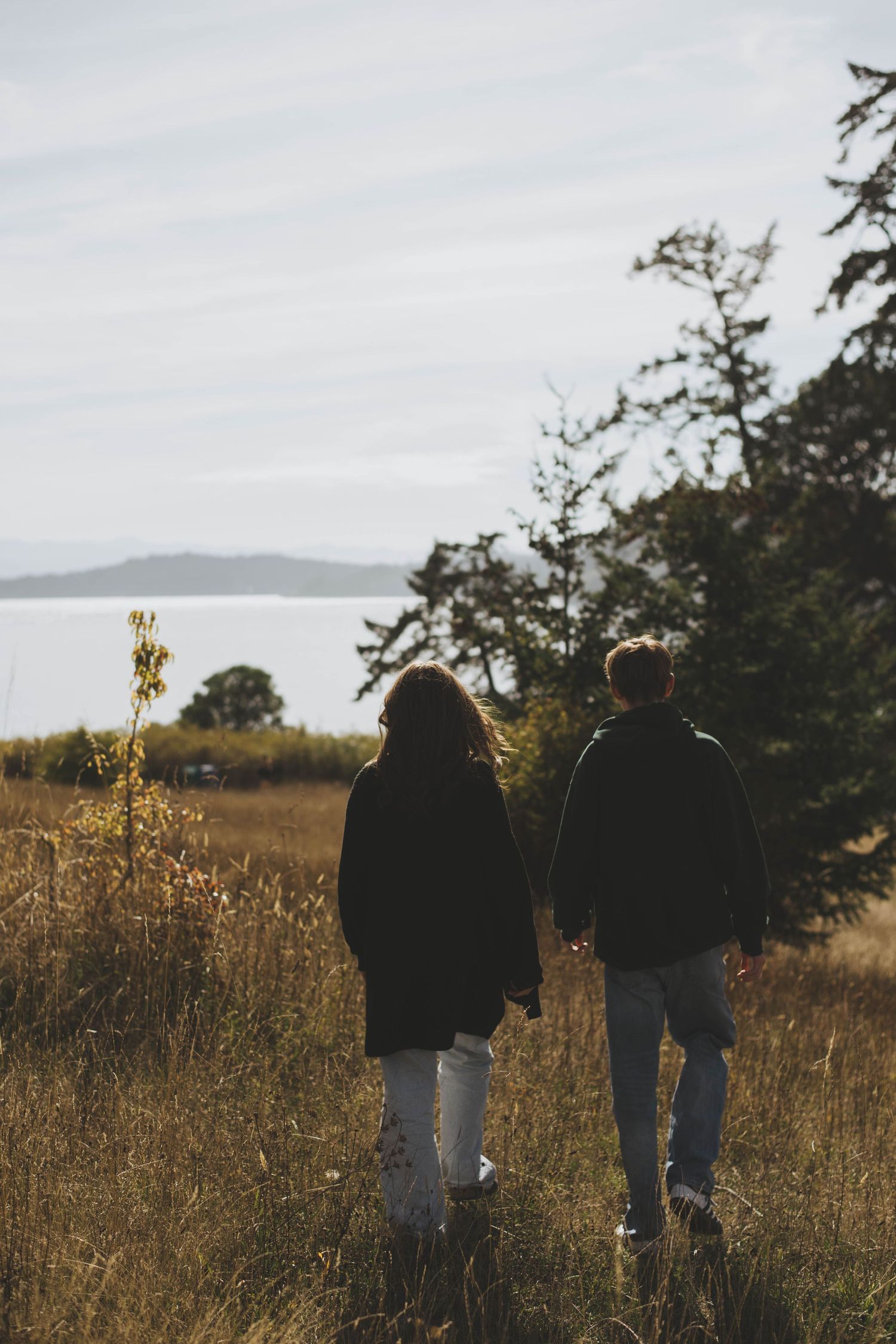 Two teenagers walk through a grassy field, with the ocean and distant mountains in view under a soft, overc