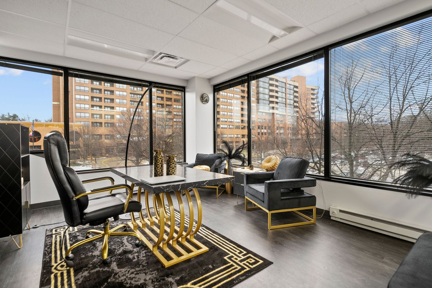 Modern office space with floor-to-ceiling windows, gold accent furniture, and dark wood floors overlooking city buildings.