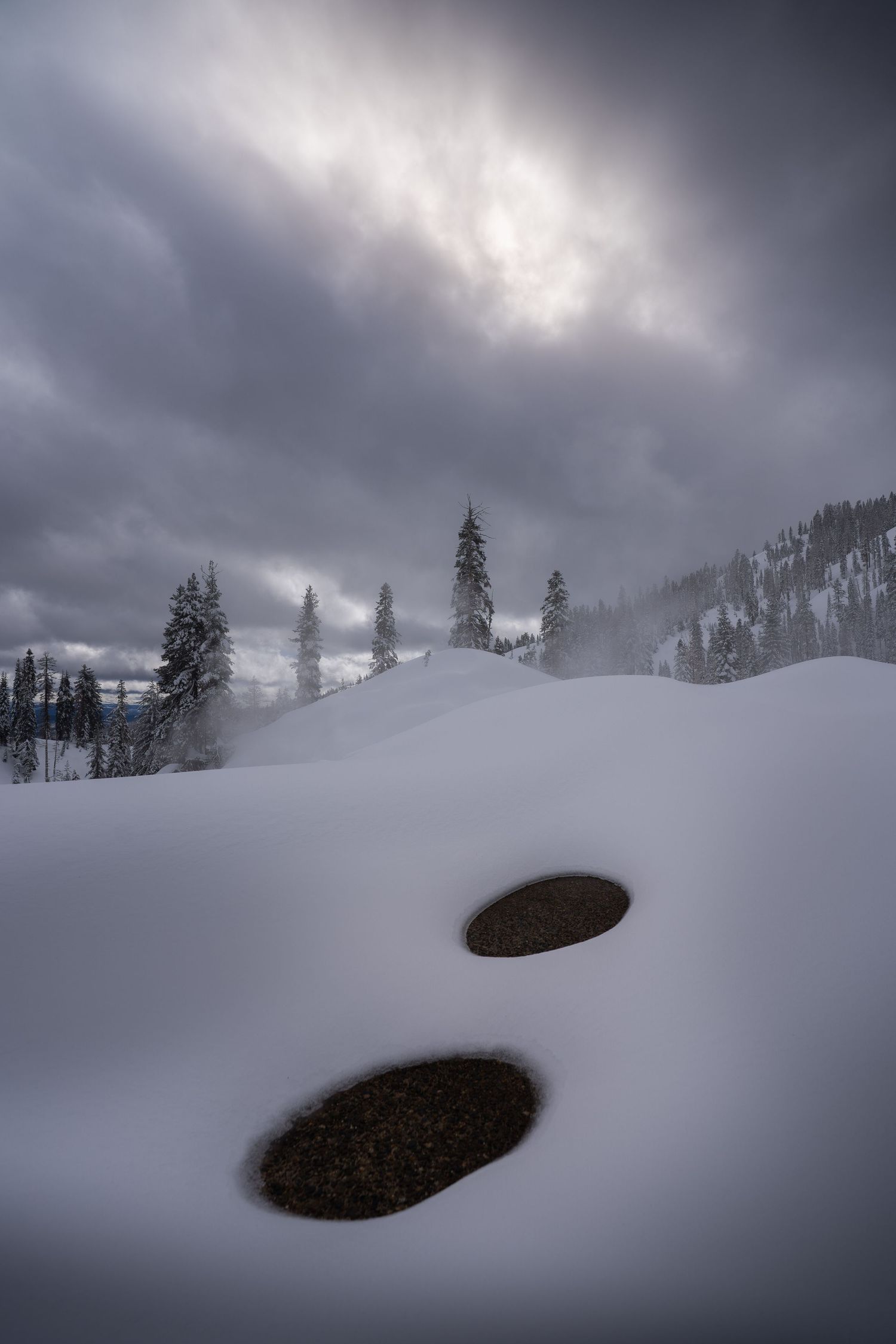 Dramatic winter landscape with two dark holes in deep snow against moody cloudy sky and evergreen trees in mountains.