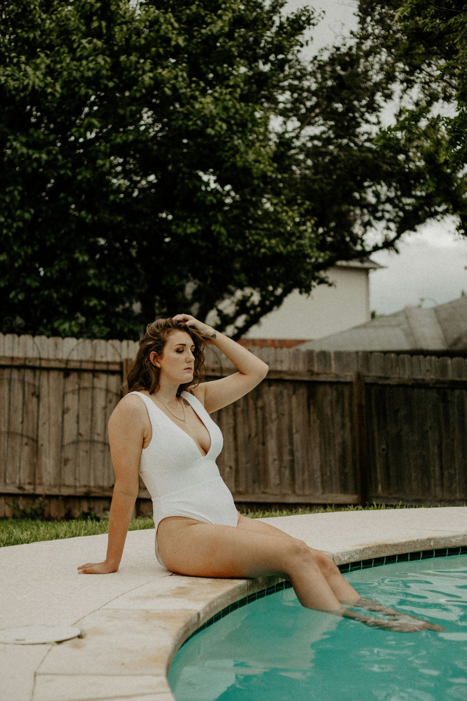 Woman in white swimsuit relaxes by turquoise swimming pool on sunny summer day near wooden fence.