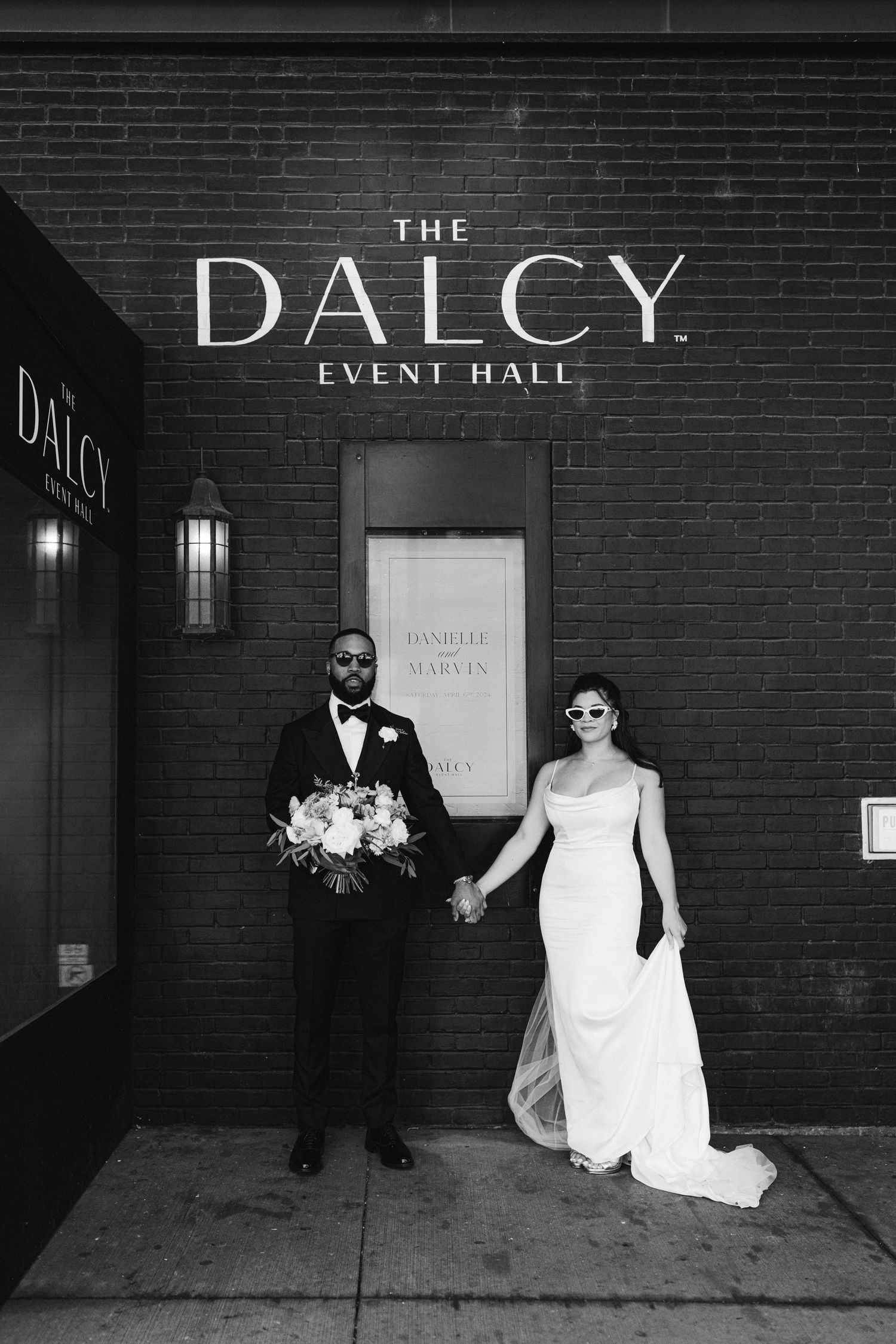 A black and white portrait of newlyweds standing in front of The Dalcy Event Hall entrance.
