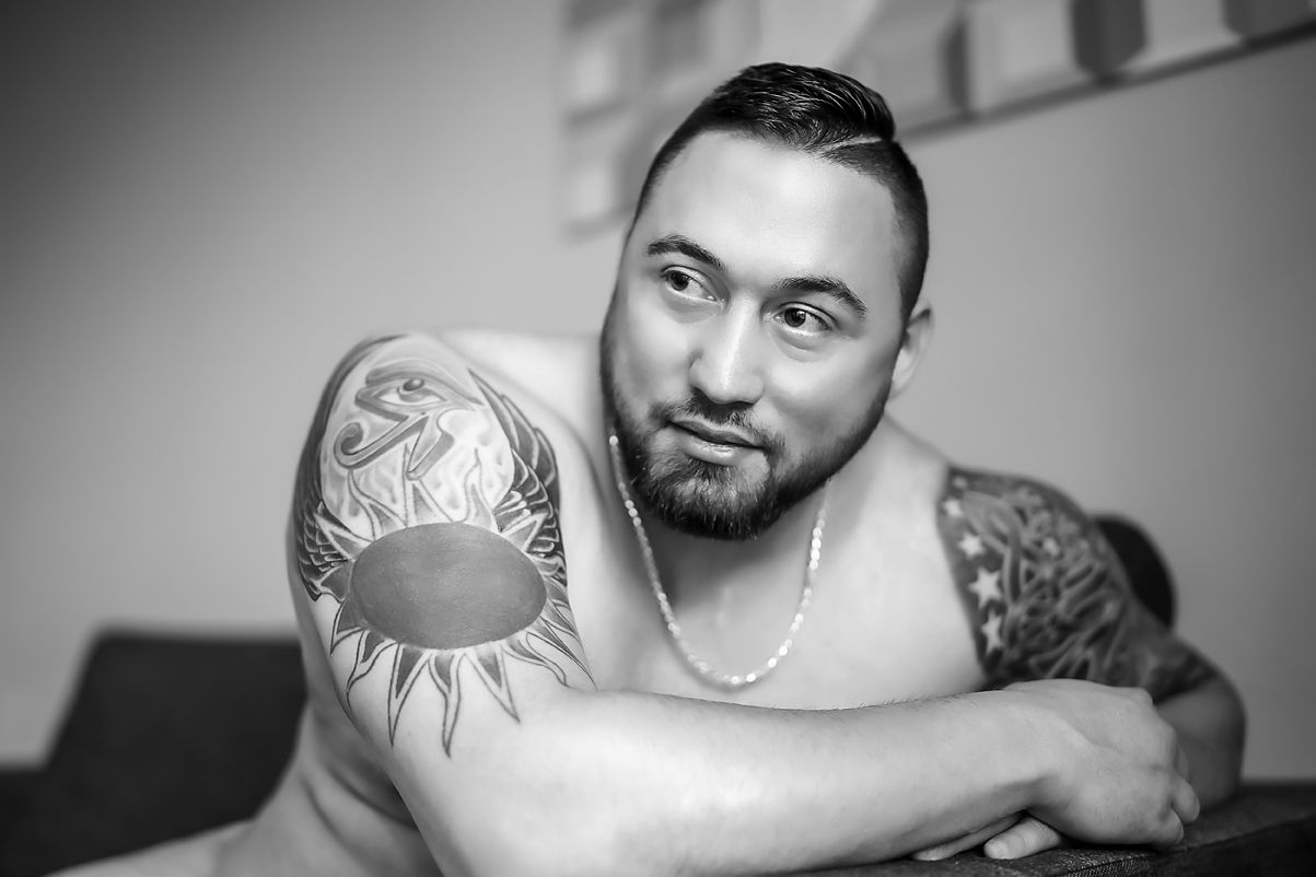 Black and white portrait of a person with tribal sun tattoo and beard posing against gray background.