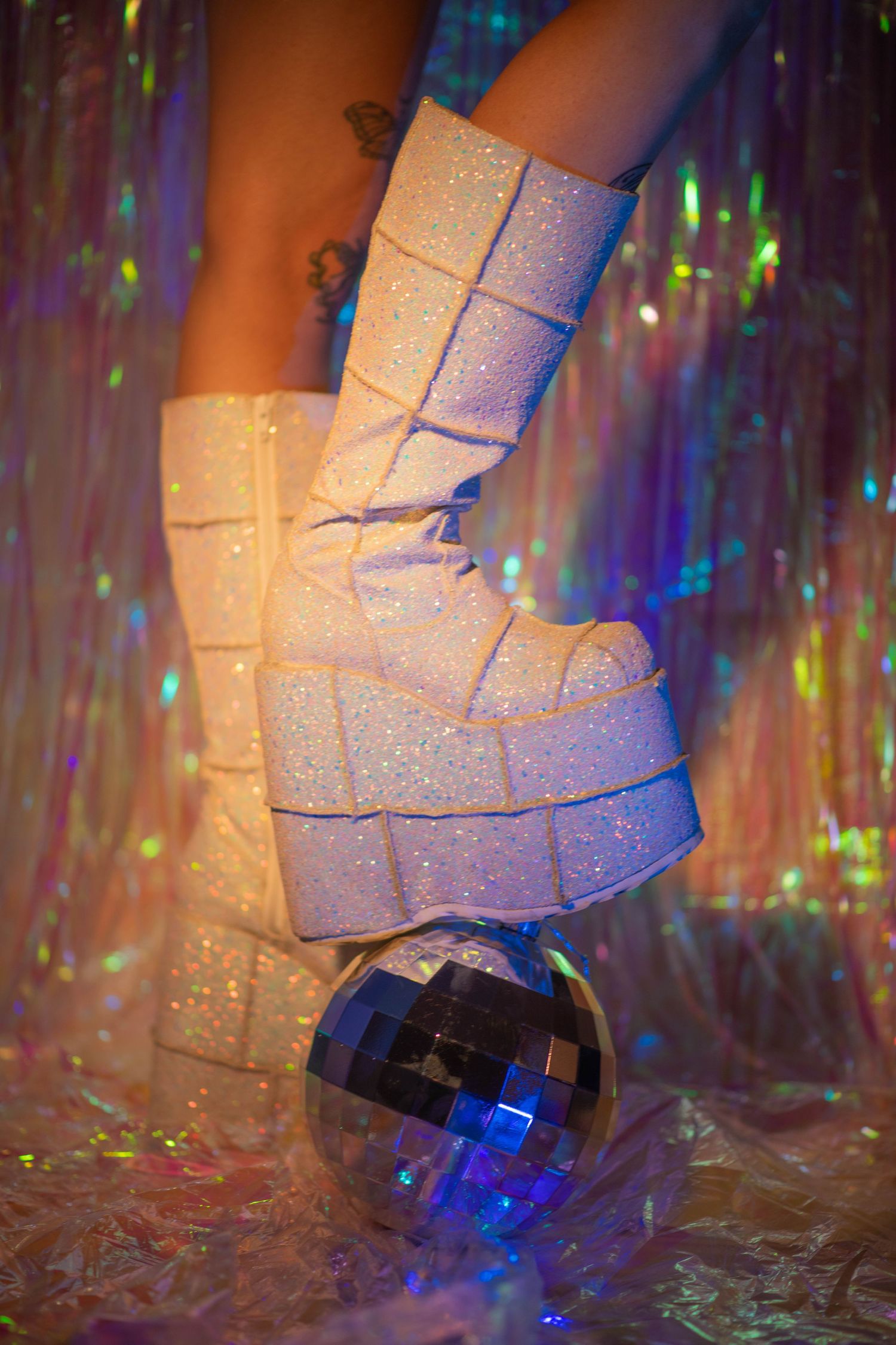 White glitter platform boot balancing on a disco ball against a holographic tinsel backdrop.