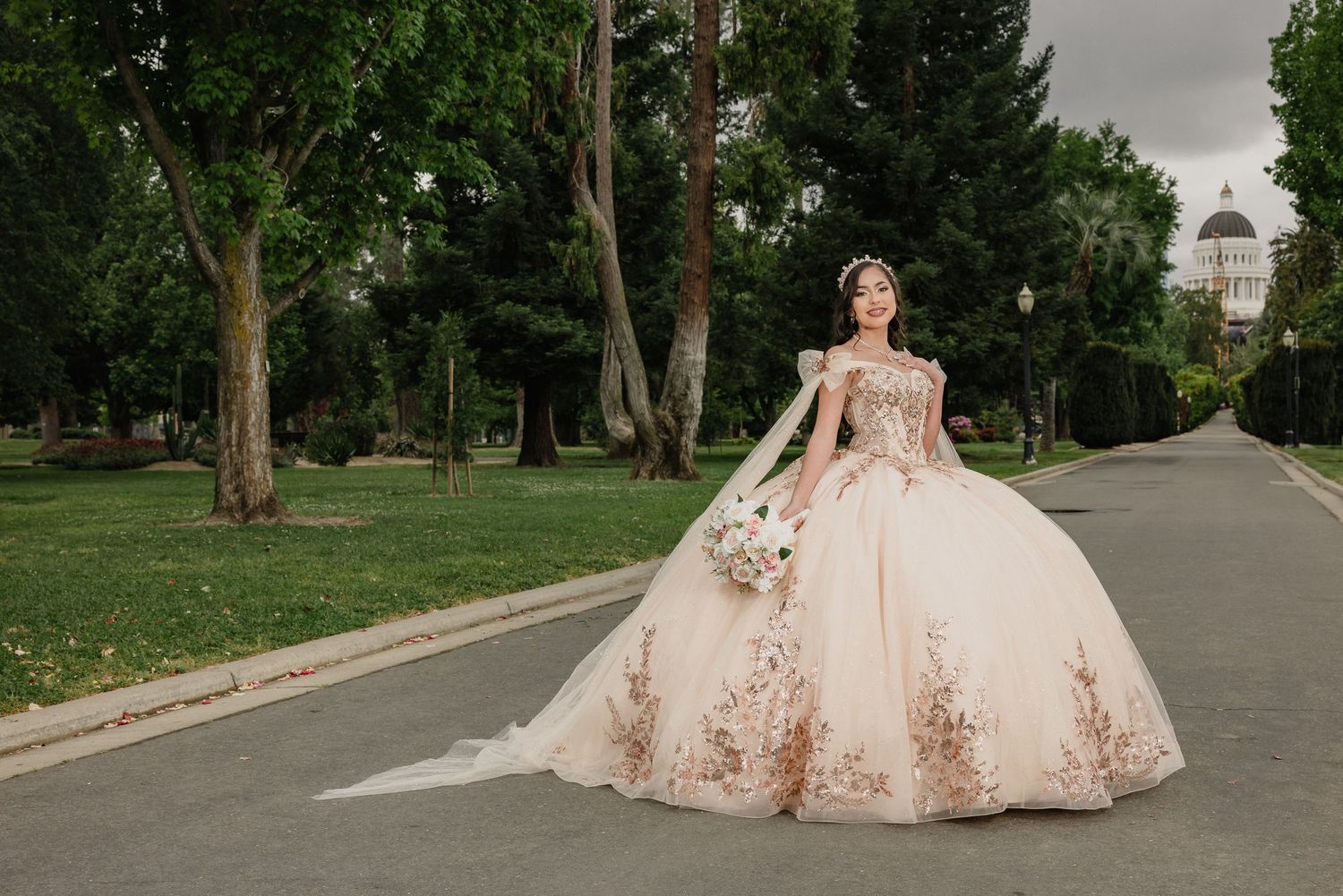 A rose gold quinceanera ball gown with floral details sparkles on a tree-lined pathway in a lush park setting.