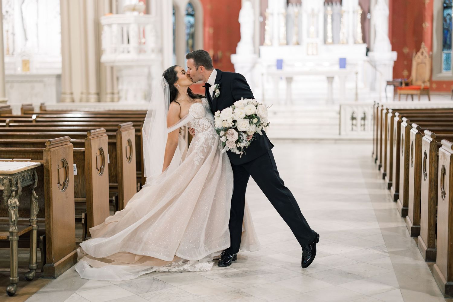 A grand cathedral interior with Gothic architecture features rows of wooden pews leading to a ornate white altar.