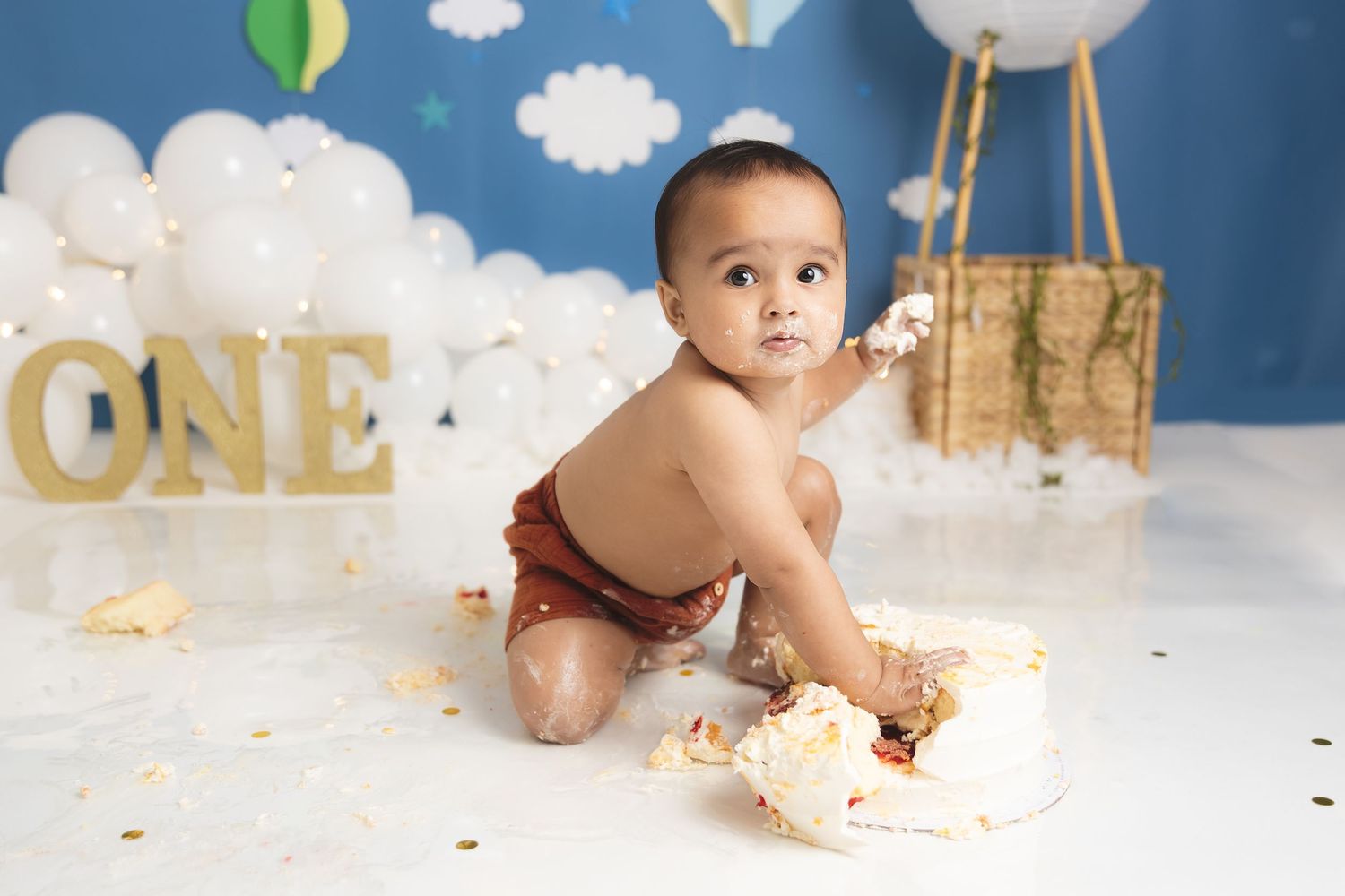 A first birthday cake smash scene with white clouds, gold 'ONE' letters, and a hot air balloon theme backdrop.