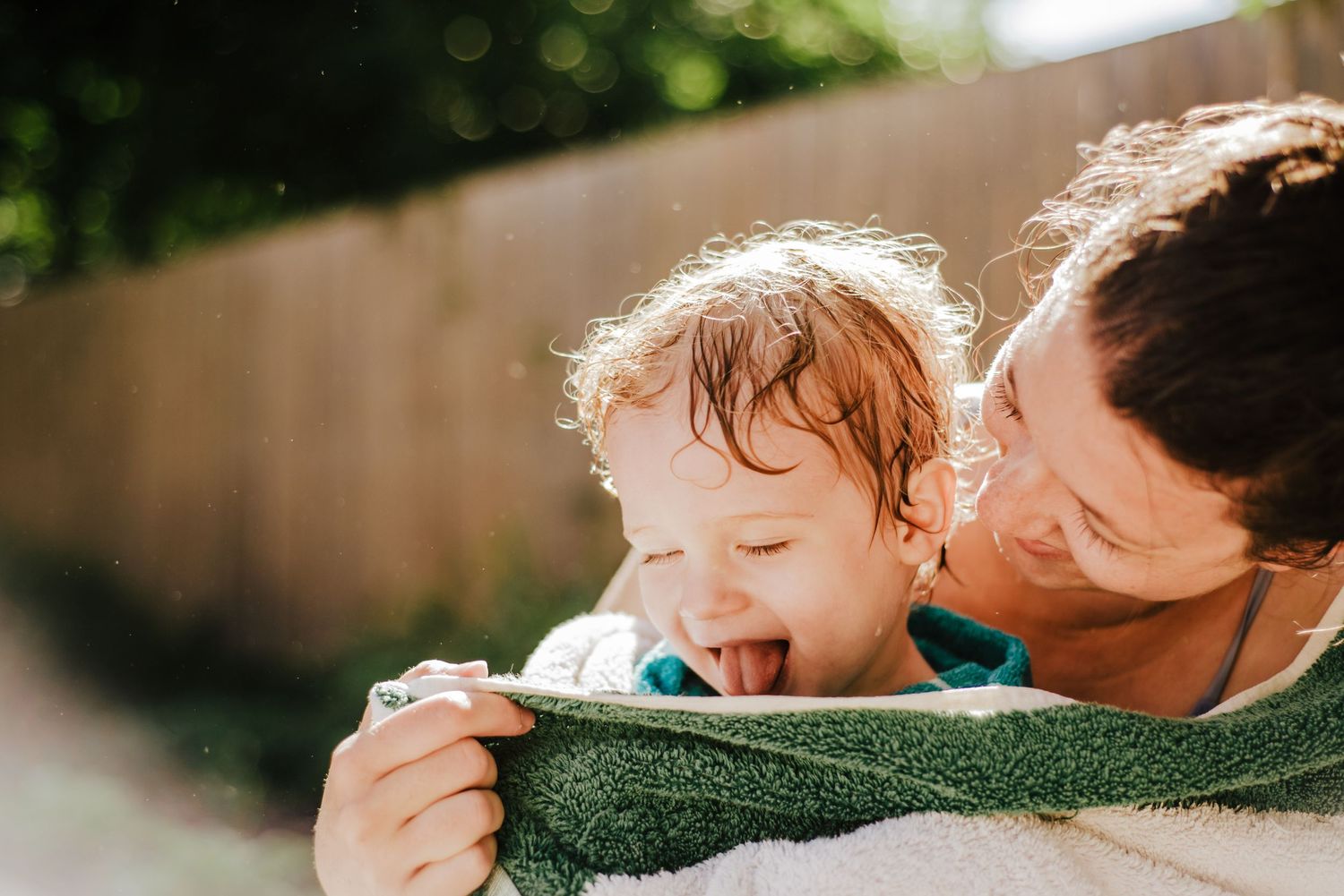 A parent and child share a tender moment wrapped in a green towel on a sunny day.
