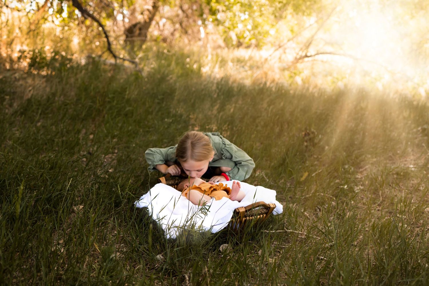 Newborn baby lay on a white blanket in a basket outside on the grass, little girl kissing babies head, sun beams behind