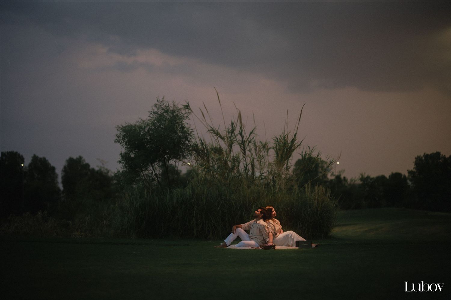 A couple sits together on a blanket in a grassy field at dusk with dramatic stormy skies overhead.