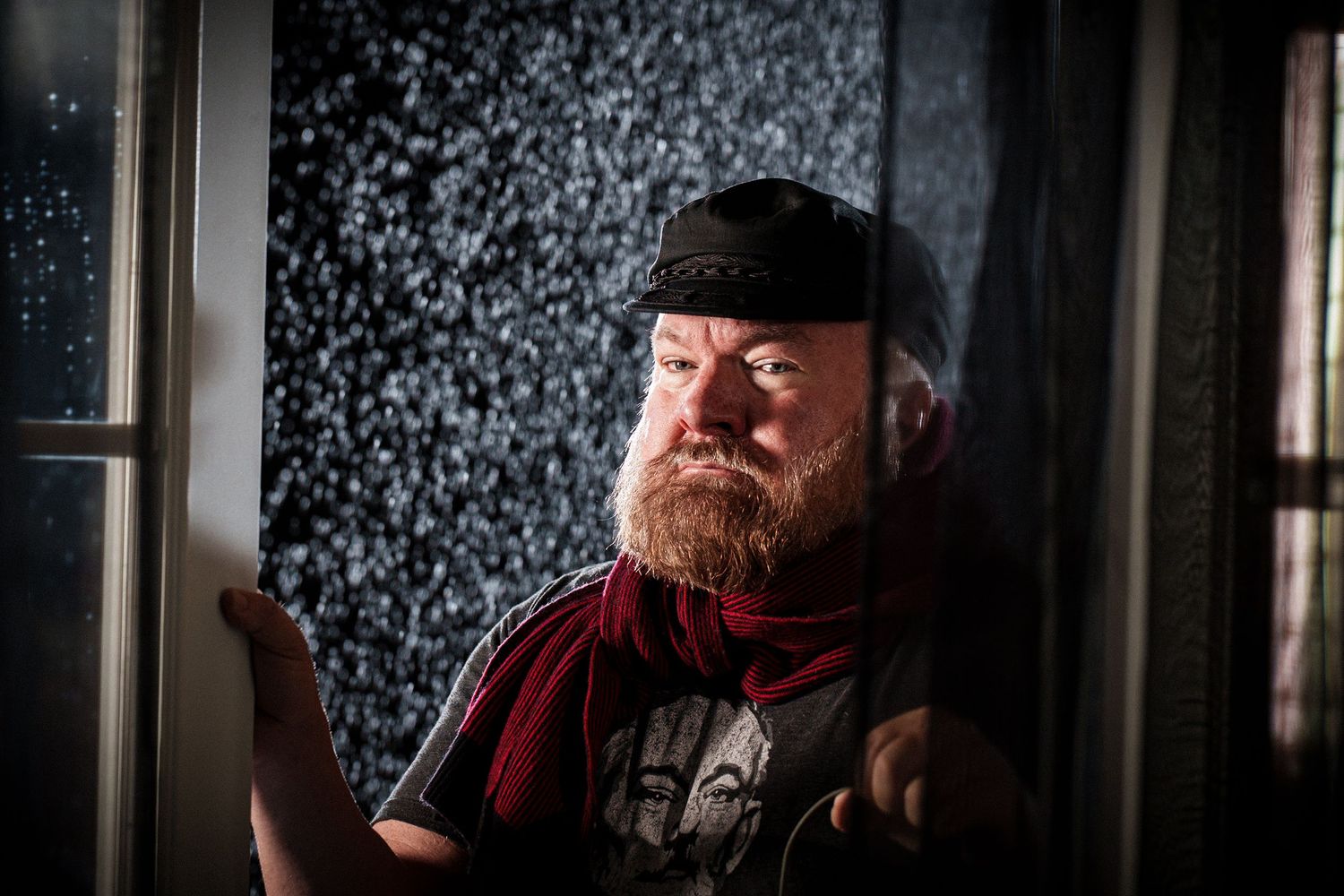 Dramatic portrait of person in black cap with red beard against textured background.