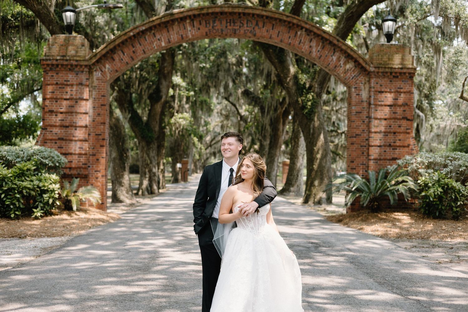 Newlyweds sharing romantic moments under a picturesque brick archway surrounded by moss-draped oak trees.