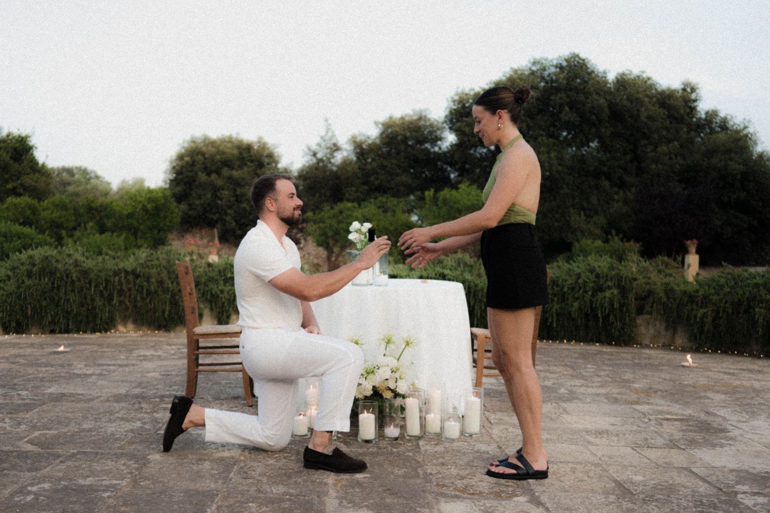 A romantic marriage proposal moment on a concrete surface decorated with candles and flowers at dusk.
