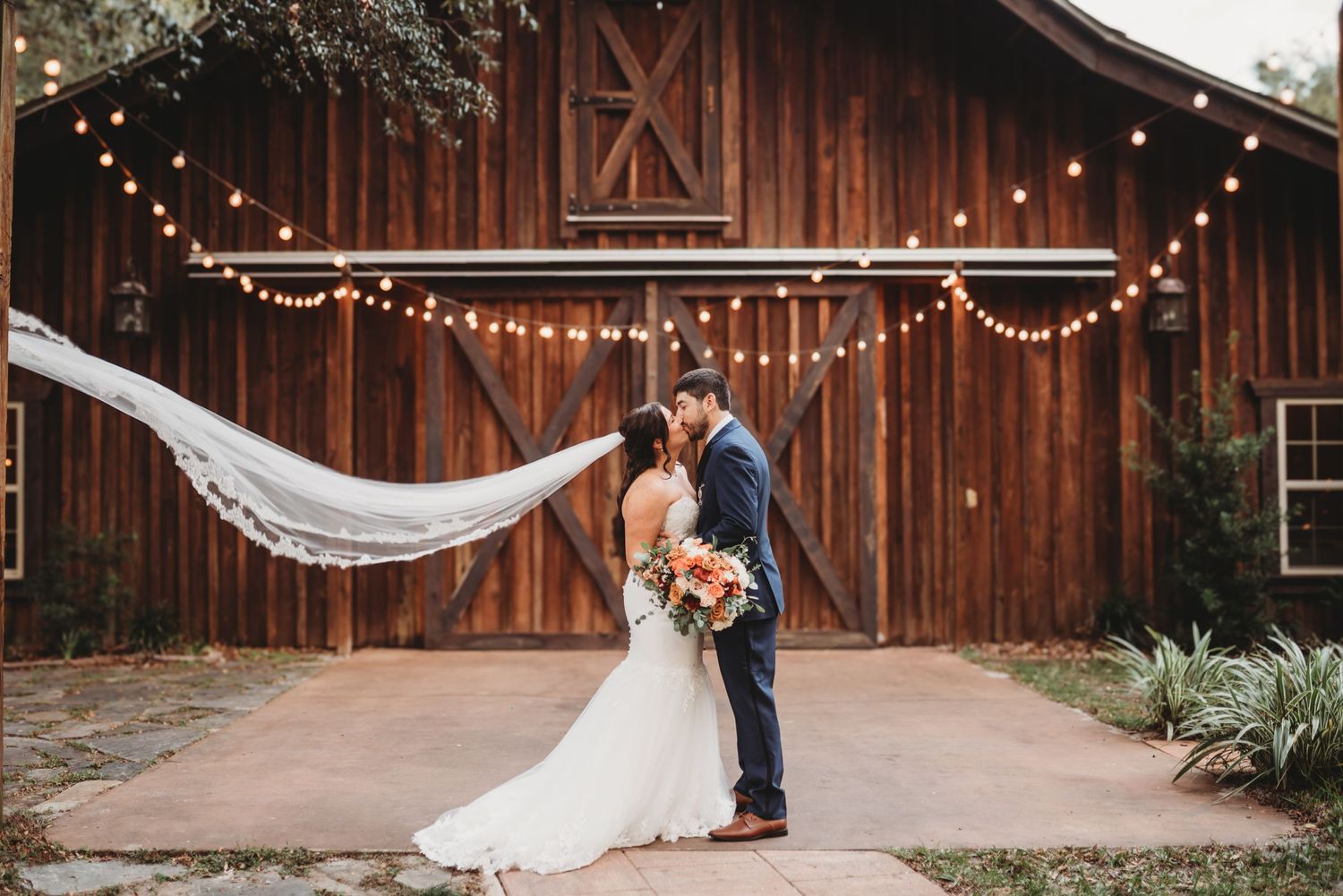 Newlyweds pose in front of rustic barn with flowing veil and string lights creating magical ambiance.