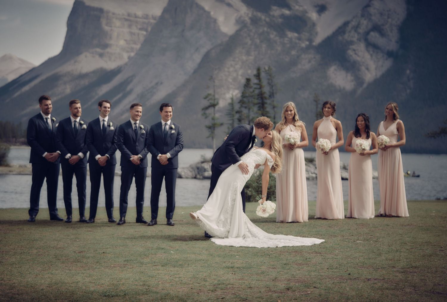 A wedding party stands against a backdrop of majestic mountain peaks and a lake at an outdoor ceremony in Banff. Wedding photographer