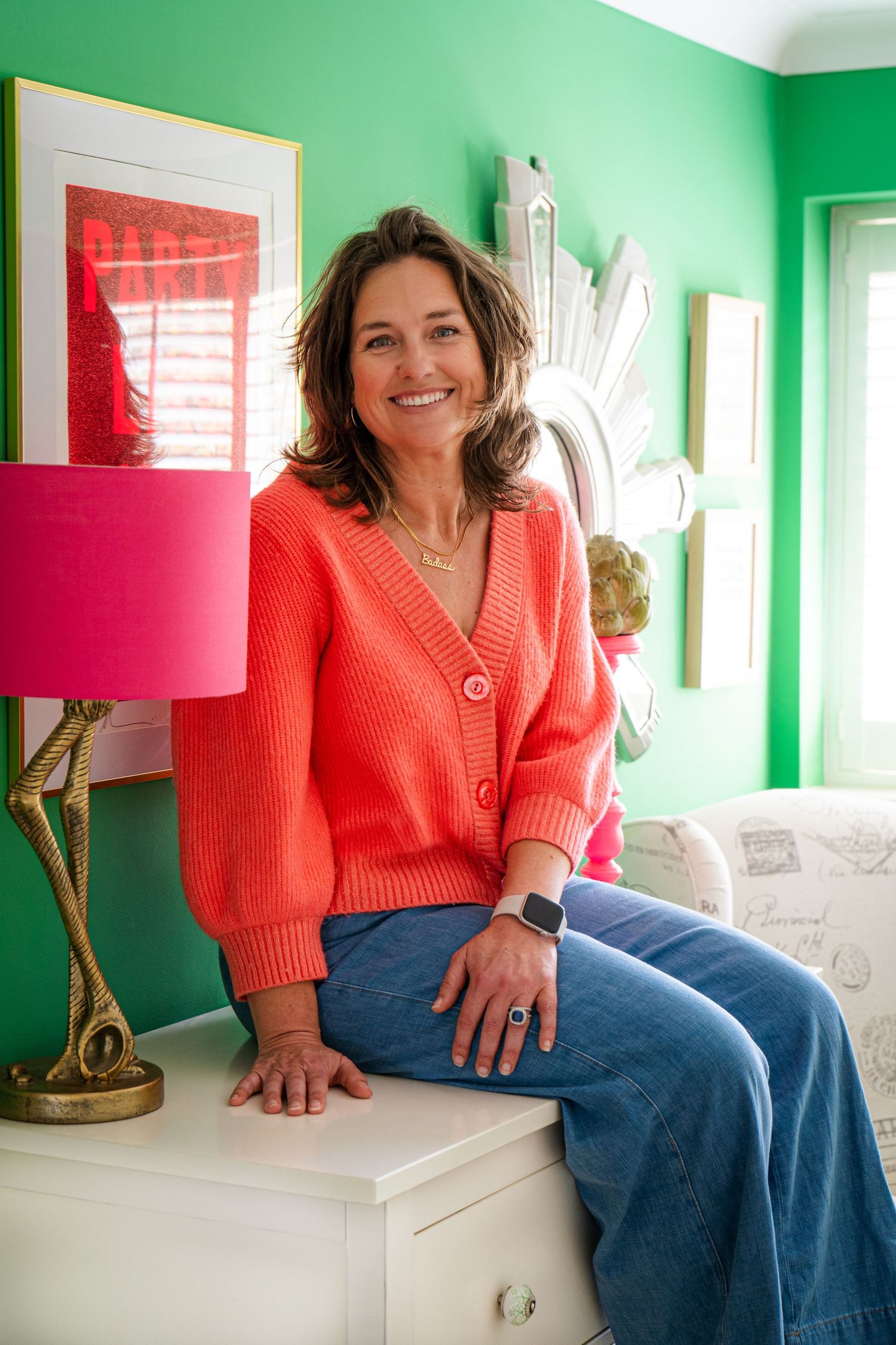 Bright interior room with green walls, pink lamp shade, and casual seating area. Female seat on unit, smiling.