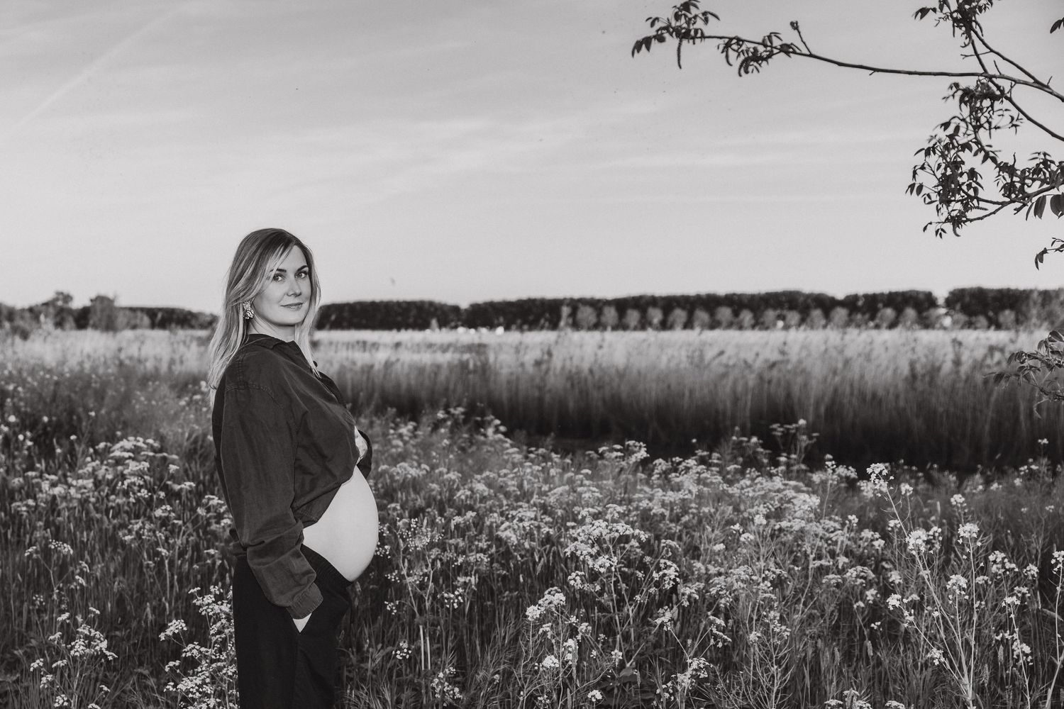 Black and white photograph of an expectant mother standing in a wildflower field with trees and sky in the background.