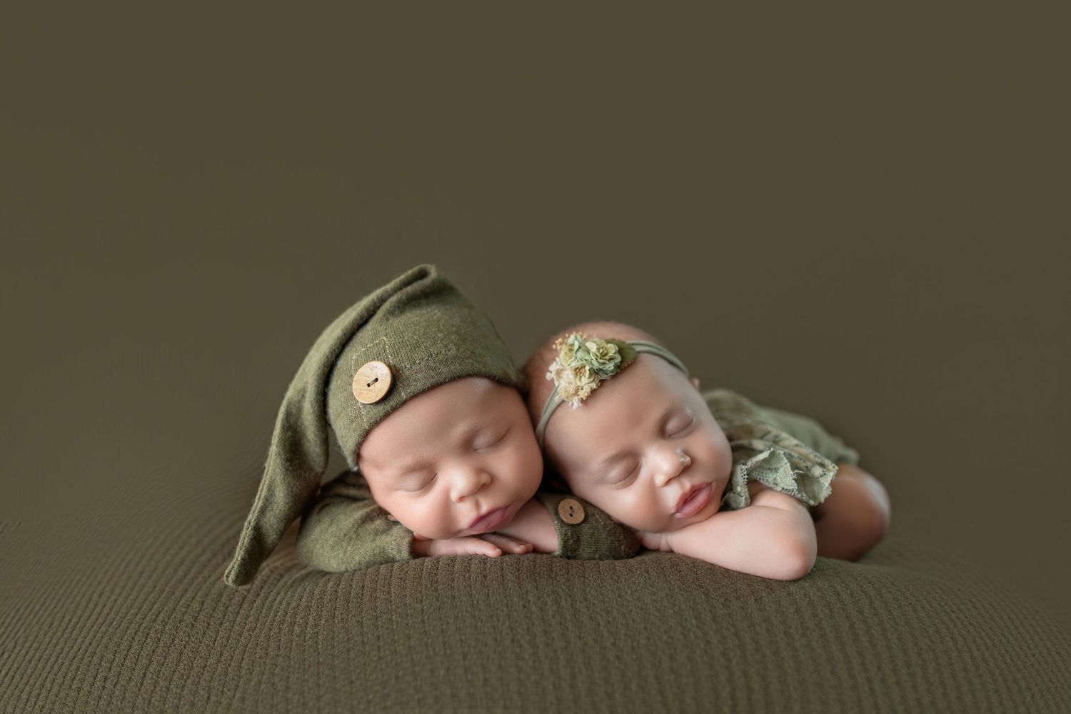Two sleeping newborn twins in matching green outfits and hats posed together on a soft green backdrop.