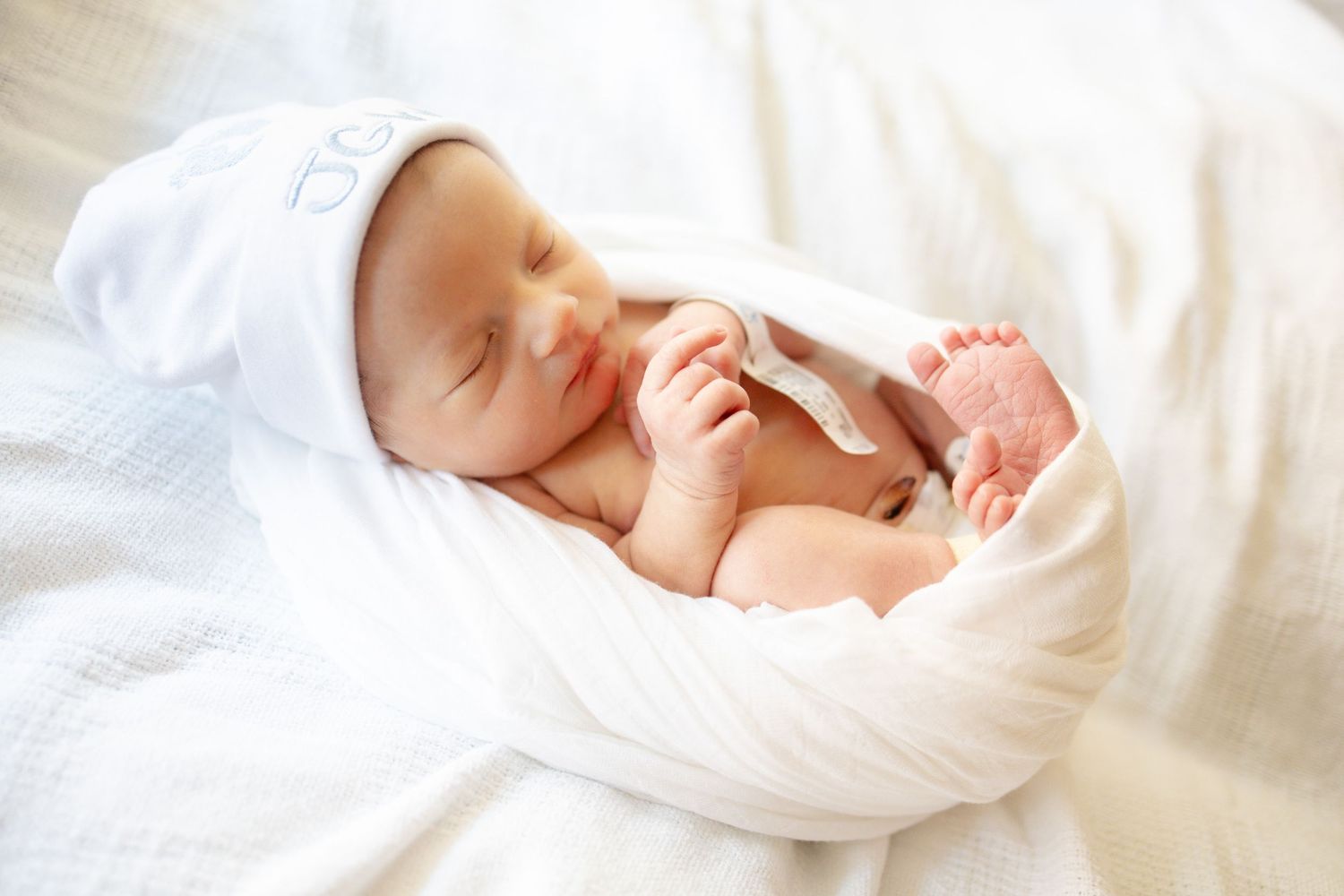 Peaceful newborn baby sleeping wrapped in soft white blanket wearing white hat on white bedding University of Tennessee Medical Center.