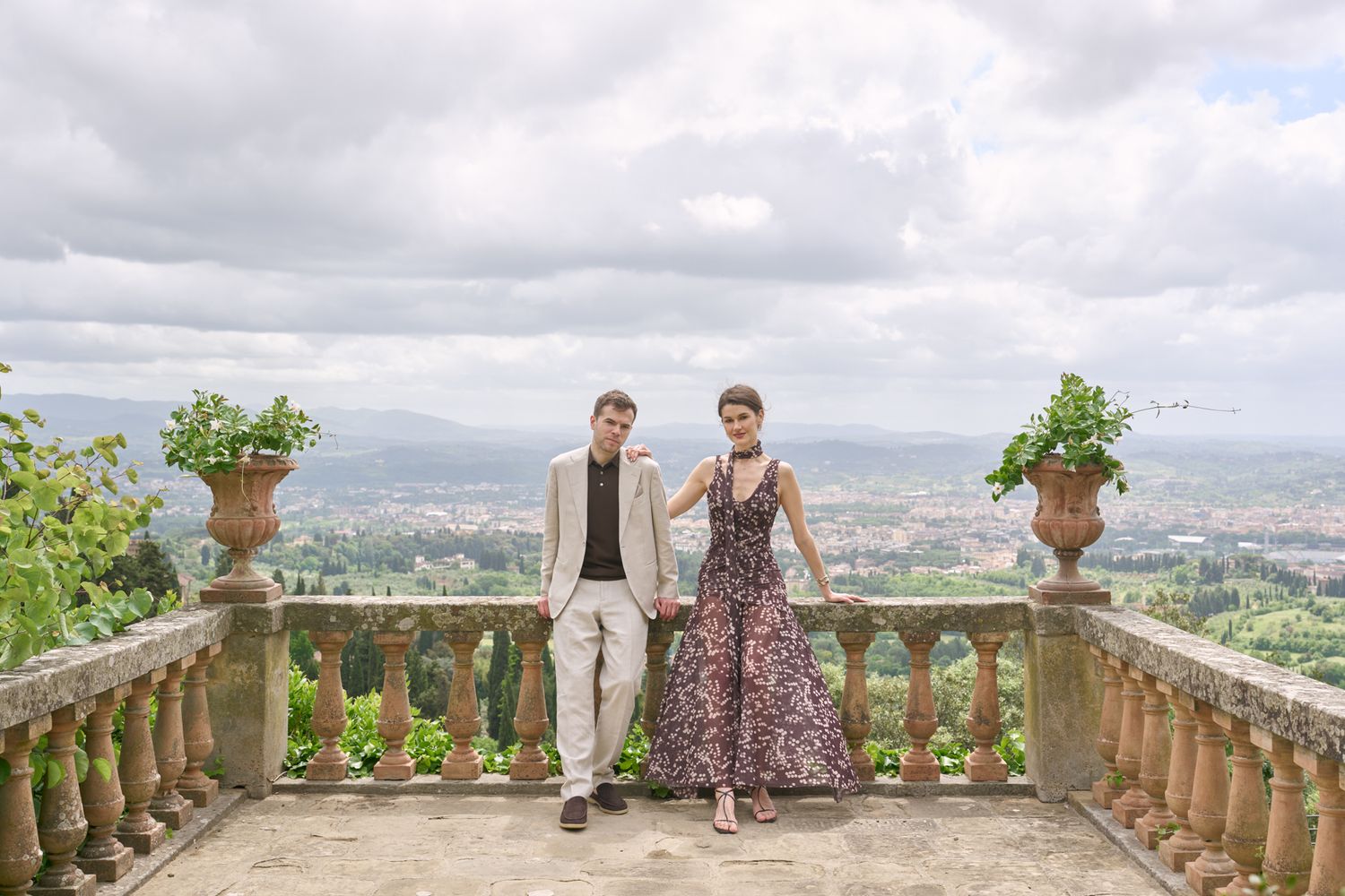 Elegant couple holding hands on a stone balcony terrace with panoramic Italian city views and cloudy sky backdrop.