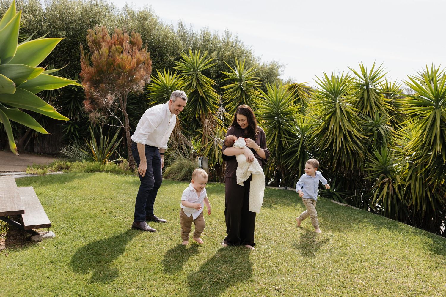 Family of five with parents, toddlers, and newborn enjoying a sunny garden surrounded by tropical plants.