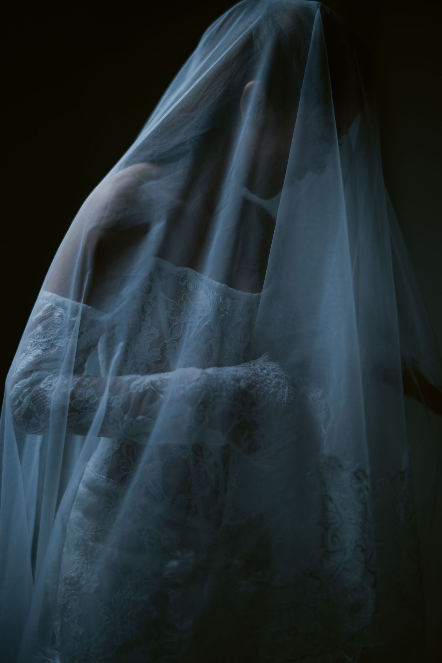 A bride in a white lace wedding dress draped with a sheer veil over her head in a dark, moody setting.