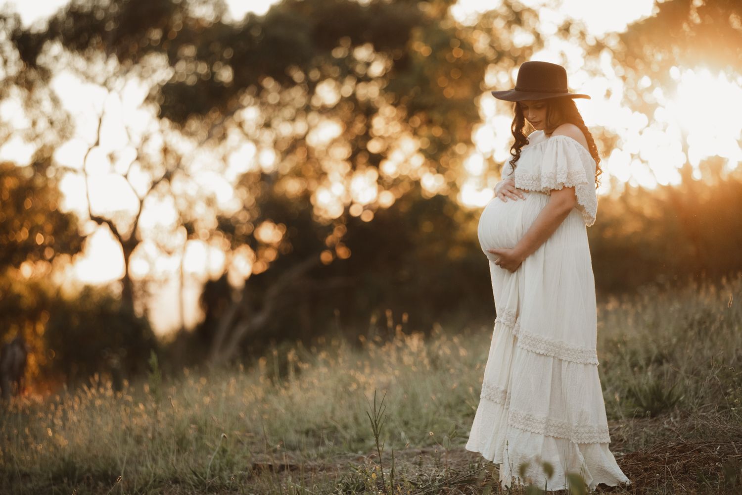 Pregnant woman in white dress and hat standing in golden sunset field, cradling baby bump in serene maternity photo.