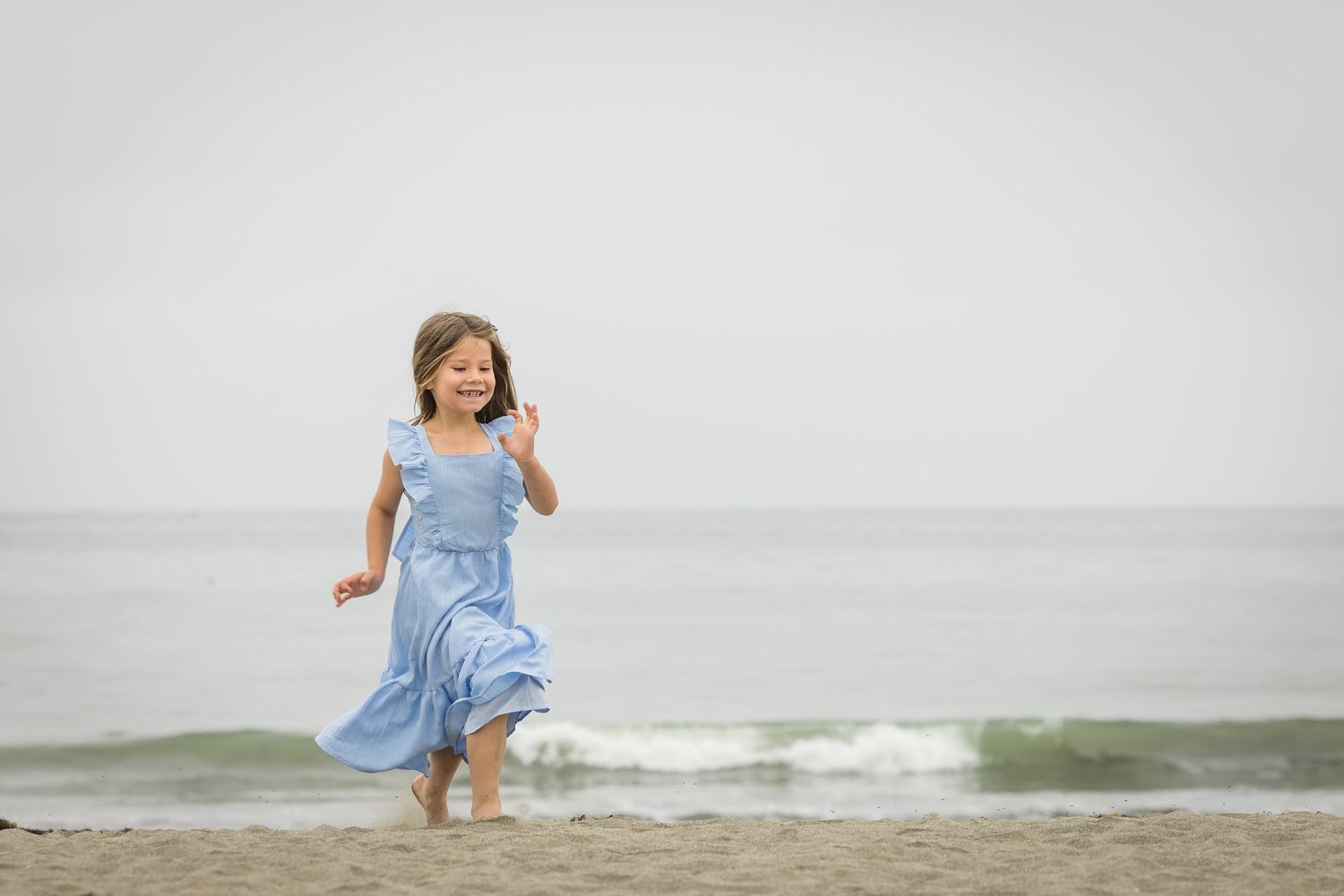 Young girl in blue dress running barefoot on sandy beach with ocean waves in background.