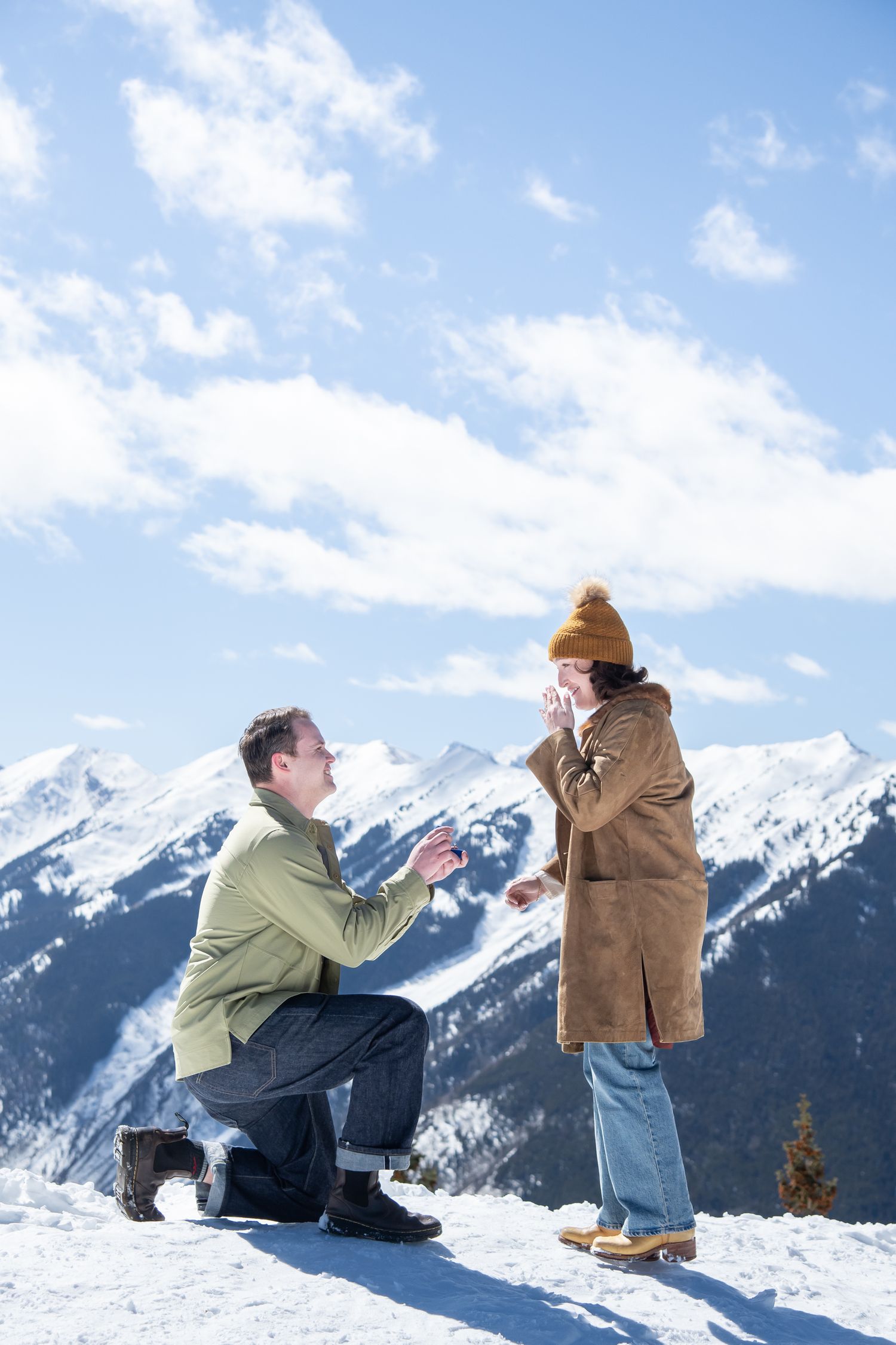 A couple in winter clothing stages a marriage proposal on a snowy mountain peak with dramatic snow-capped peaks in background in Aspen CO.