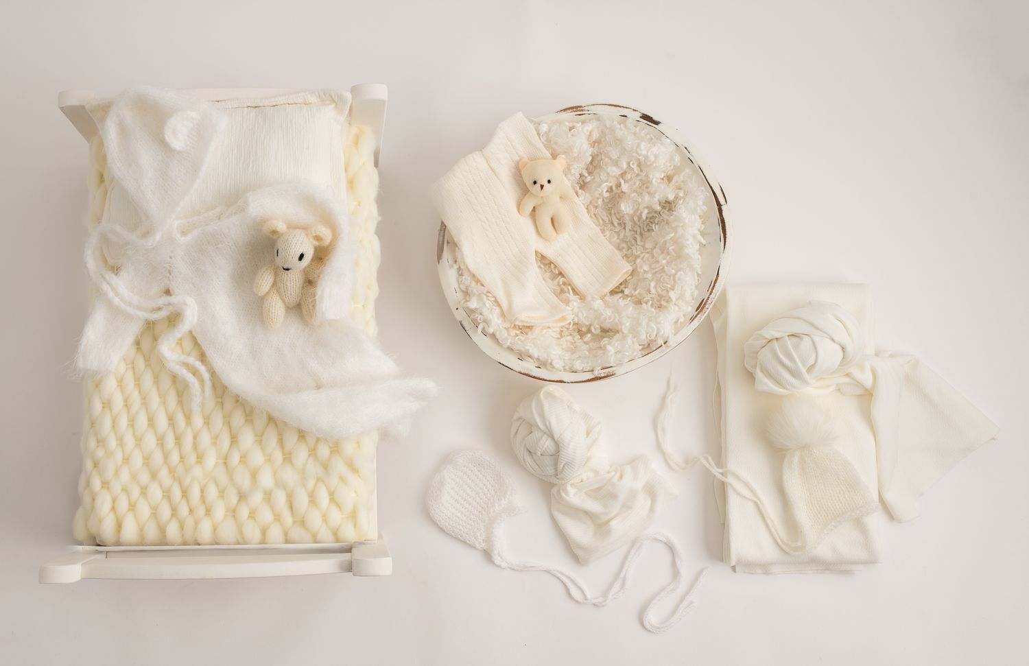 Flat lay of white and cream newborn photography props including a tiny bed, bowl, and outfit set