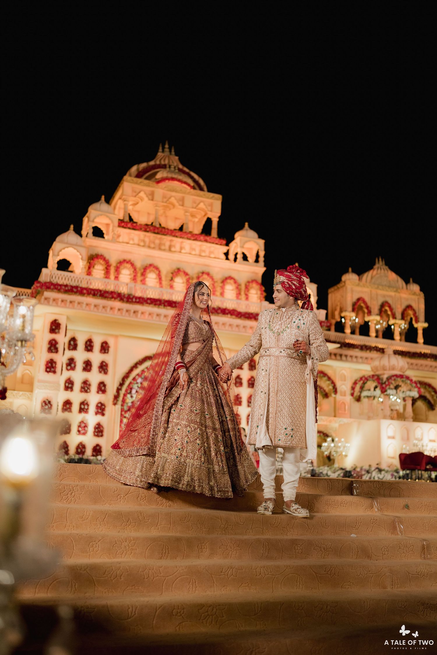 Indian bride and groom in traditional attire hold hands on steps of illuminated palace at night.