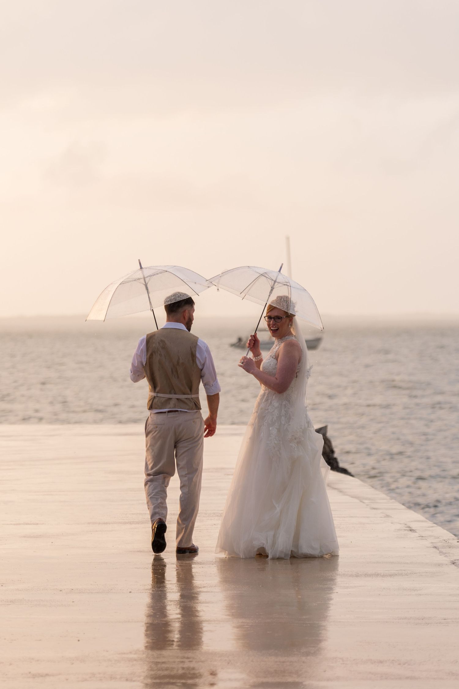 Bride and groom walking on a wet pier in the rain holding clear umbrellas with the ocean in the background.