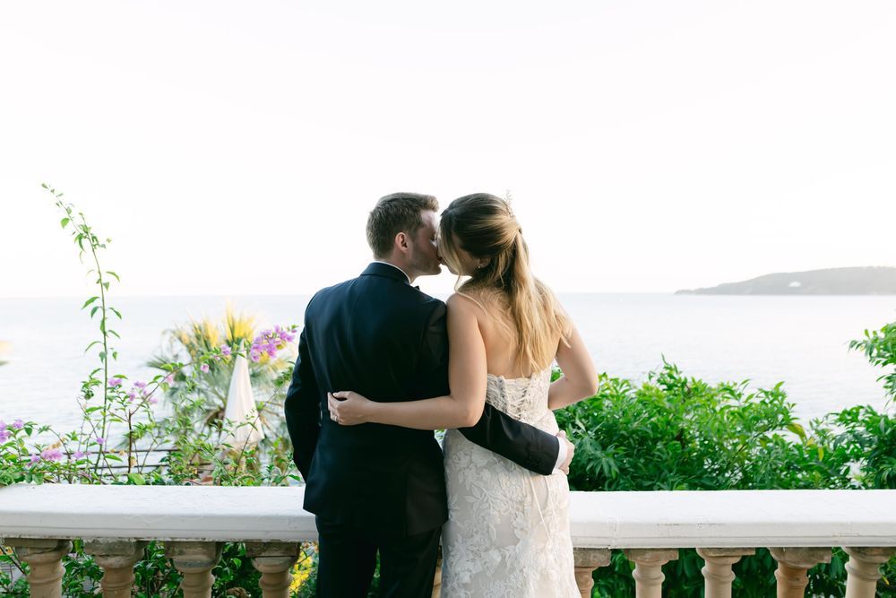 Bride and groom share a romantic kiss on a balcony overlooking the ocean, surrounded by lush greenery and colorful flowers.