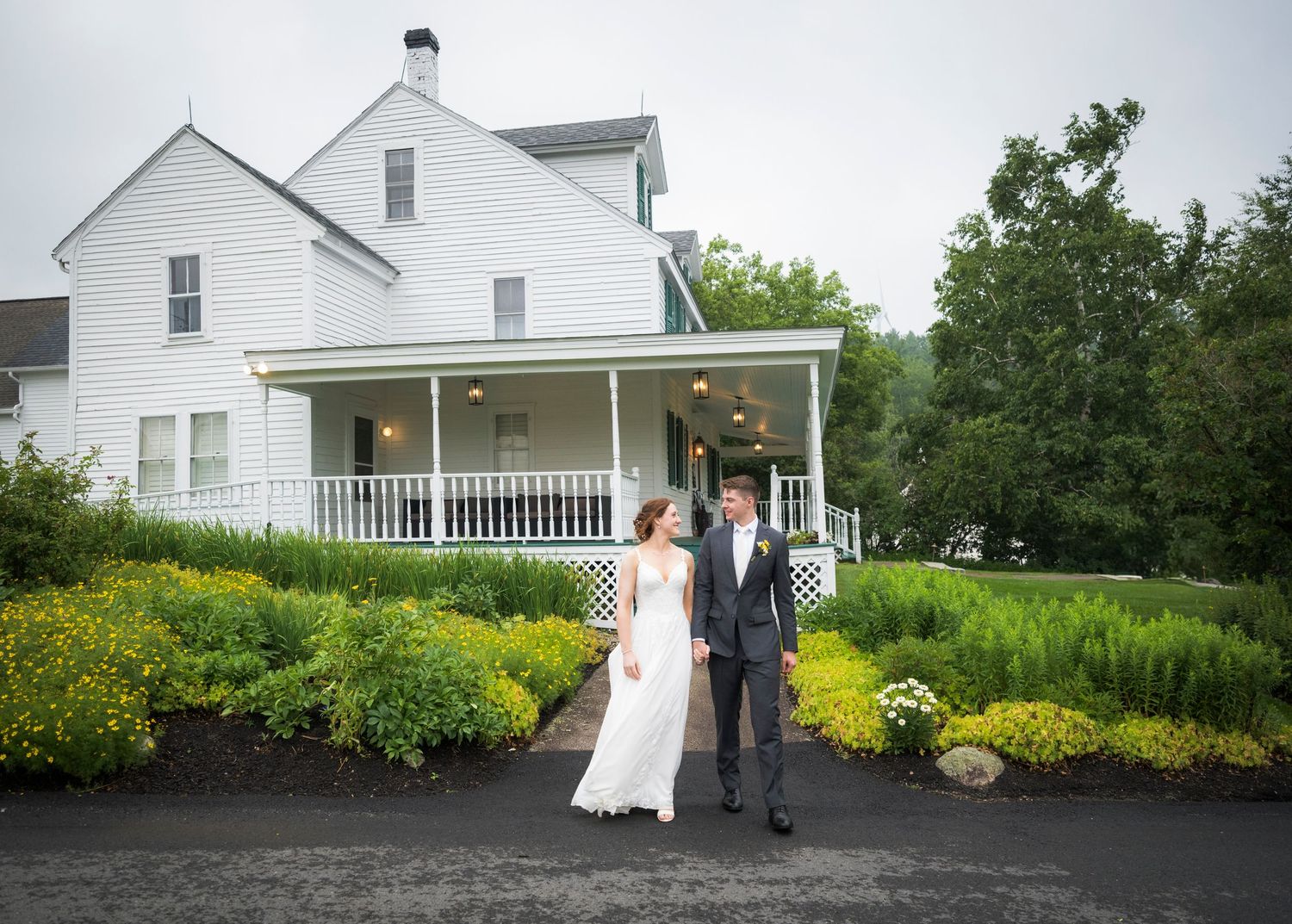 Bride and groom walk hand in hand in front of a charming white farmhouse wedding venue surrounded by lush gardens.