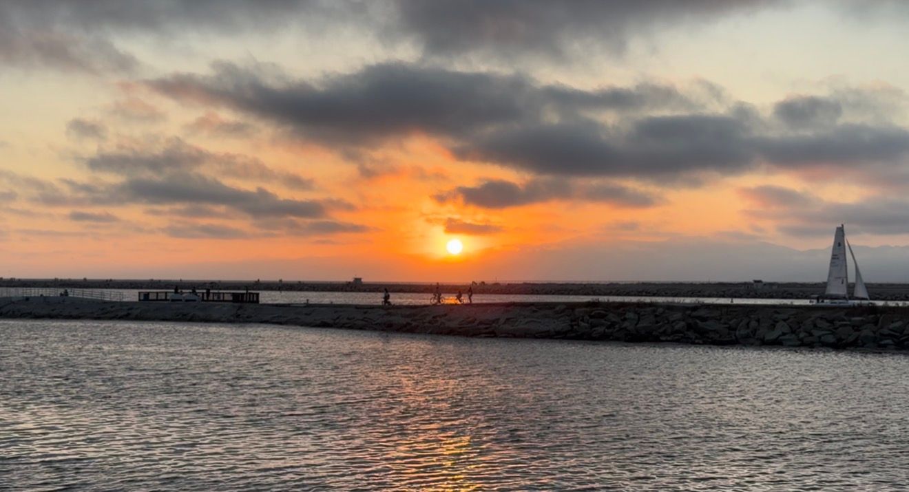 Stunning sunset over calm harbor waters with sailboat silhouette and rocky breakwater pier under dramatic cloudy sky.