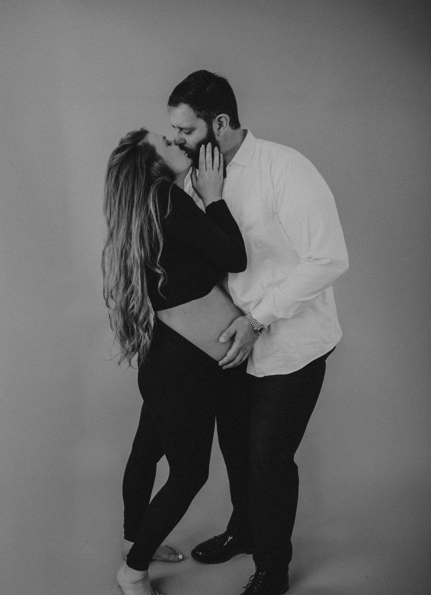 Black and white maternity photo of a couple kissing, man tenderly holding pregnant woman's belly in a studio setting.
