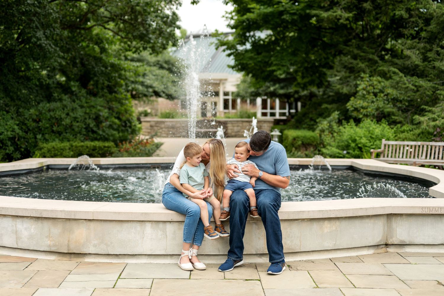 Family of four sitting by a fountain in a lush garden, parents lovingly interacting with two young children.