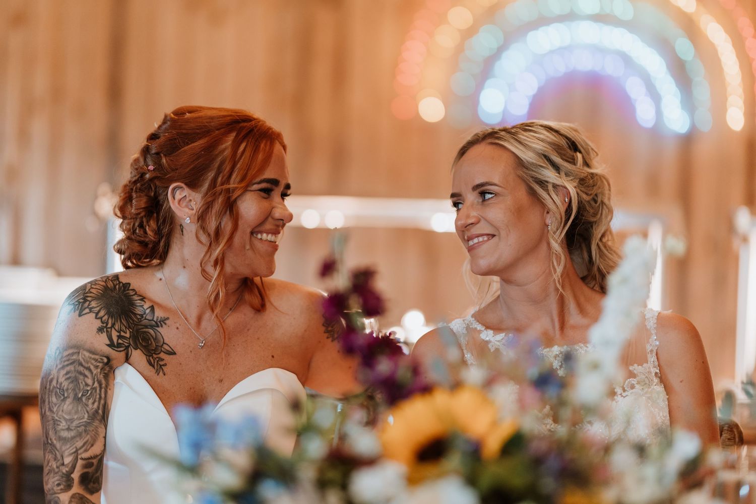 Two brides in white wedding gowns smile at each other at a decorated reception table with colorful floral arrangements.