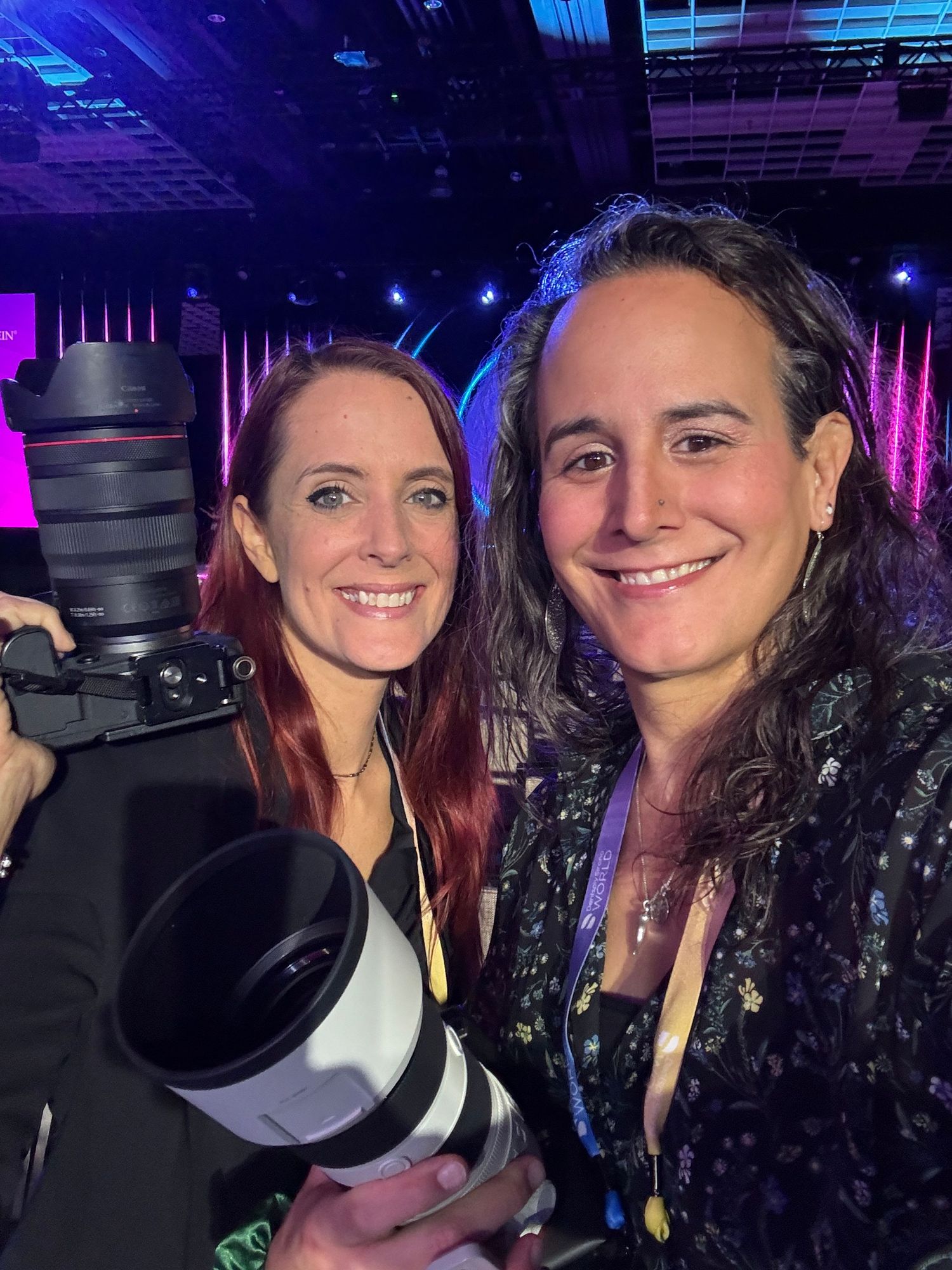 Two women smiling at a event, one holding a professional camera with a large telephoto lens, colorful stage lighting behind them.