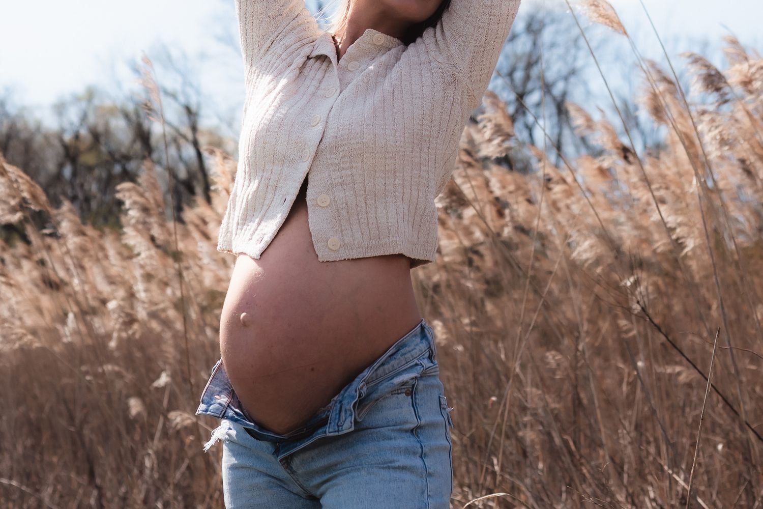 Pregnant woman in crop top and jeans standing in golden field, showcasing baby bump outdoors.