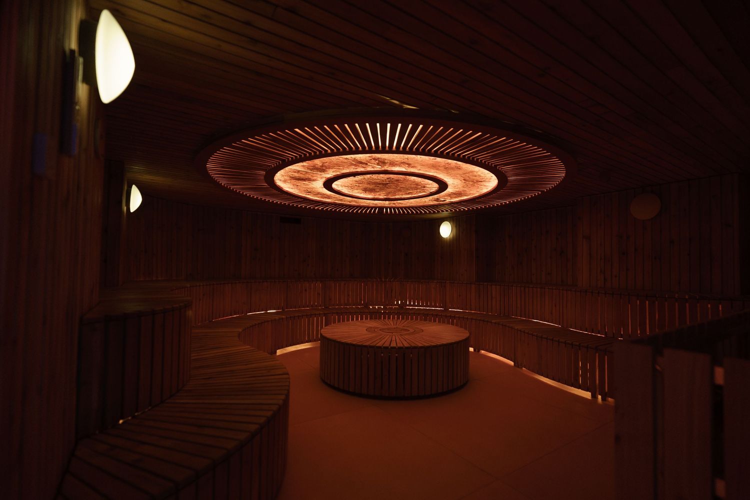 Warm dimly lit sauna room with wooden benches, circular ceiling light fixture, and a central round wooden structure.