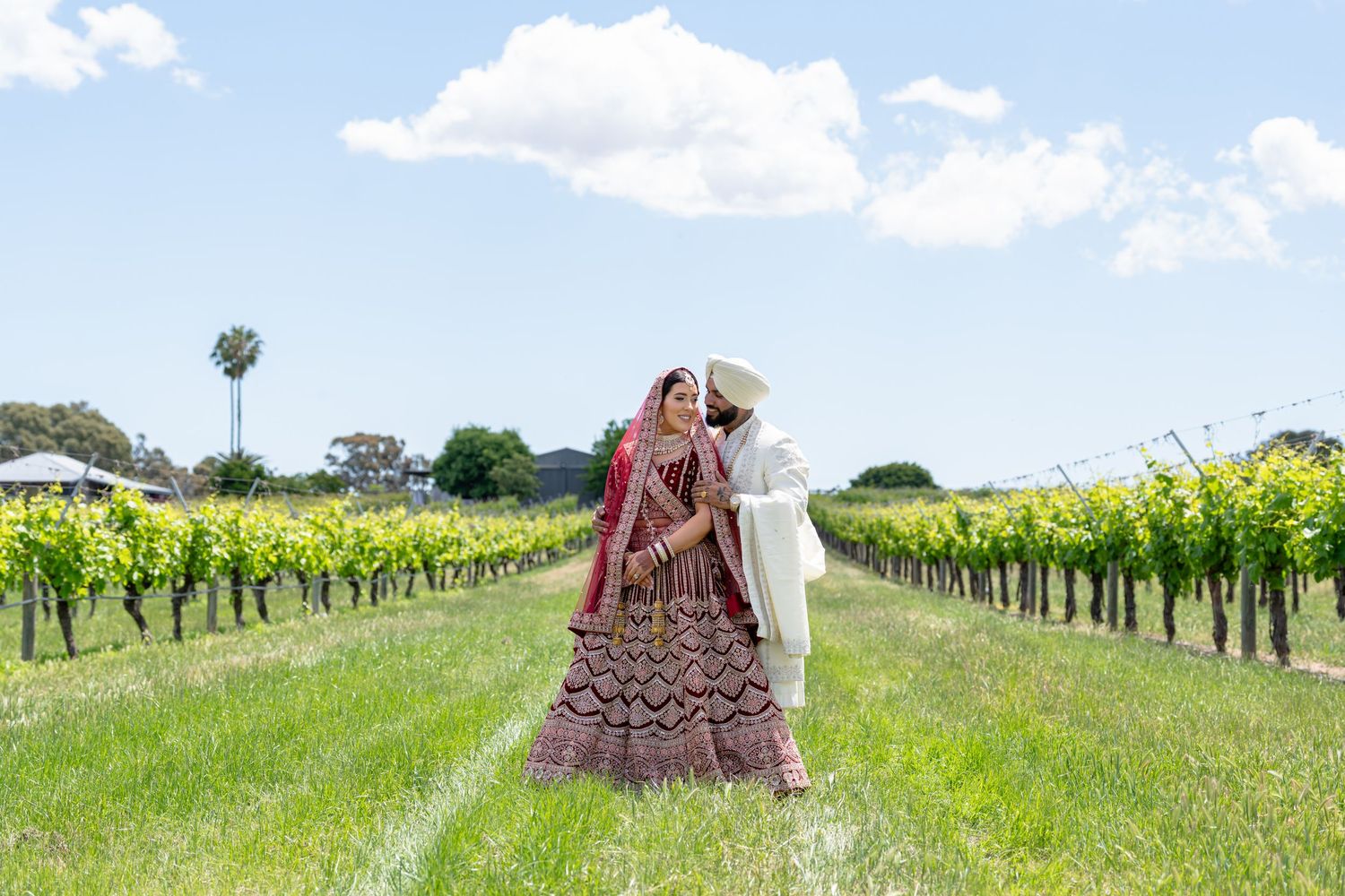 Indian bride and groom pose beside a classic white Rolls-Royce adorned with red ribbons on their wedding day.